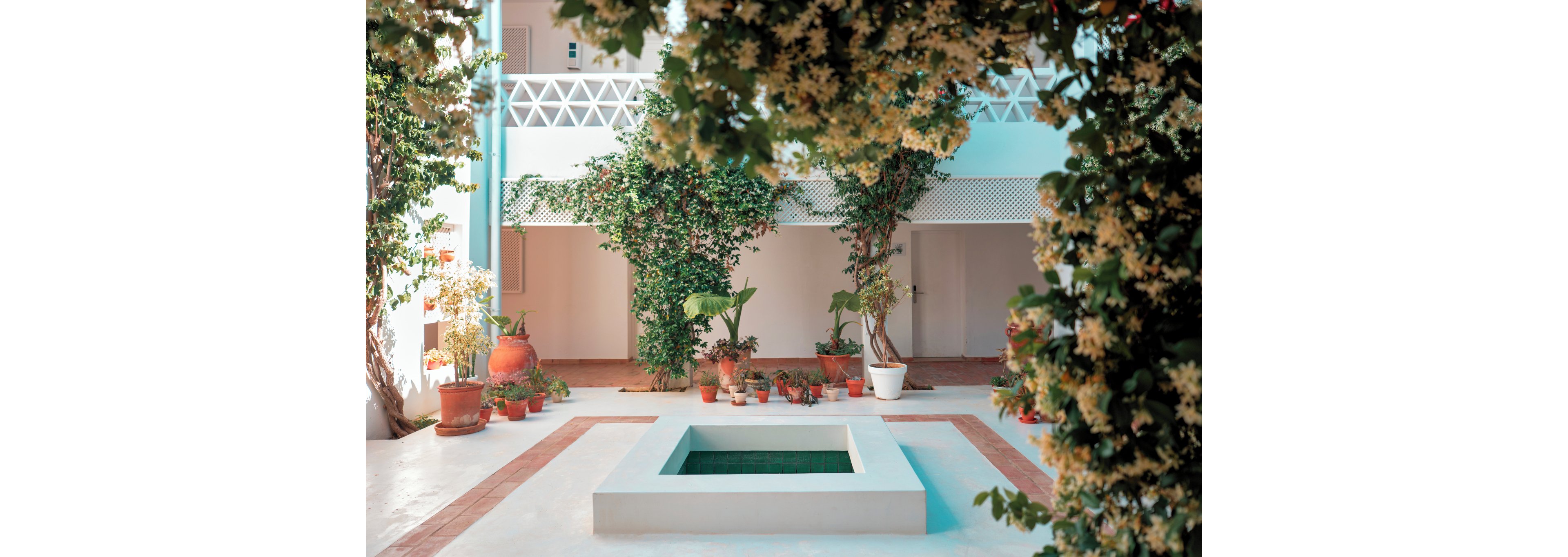 A bright courtyard features a small square fountain at its center, surrounded by potted plants. Climbing vines and flowering greenery frame the scene, creating a lush, intimate atmosphere. Soft pastel walls and geometric balcony railings add a modern yet Mediterranean feel. Warm sunlight enhances the contrast between the white floor and the vibrant foliage.