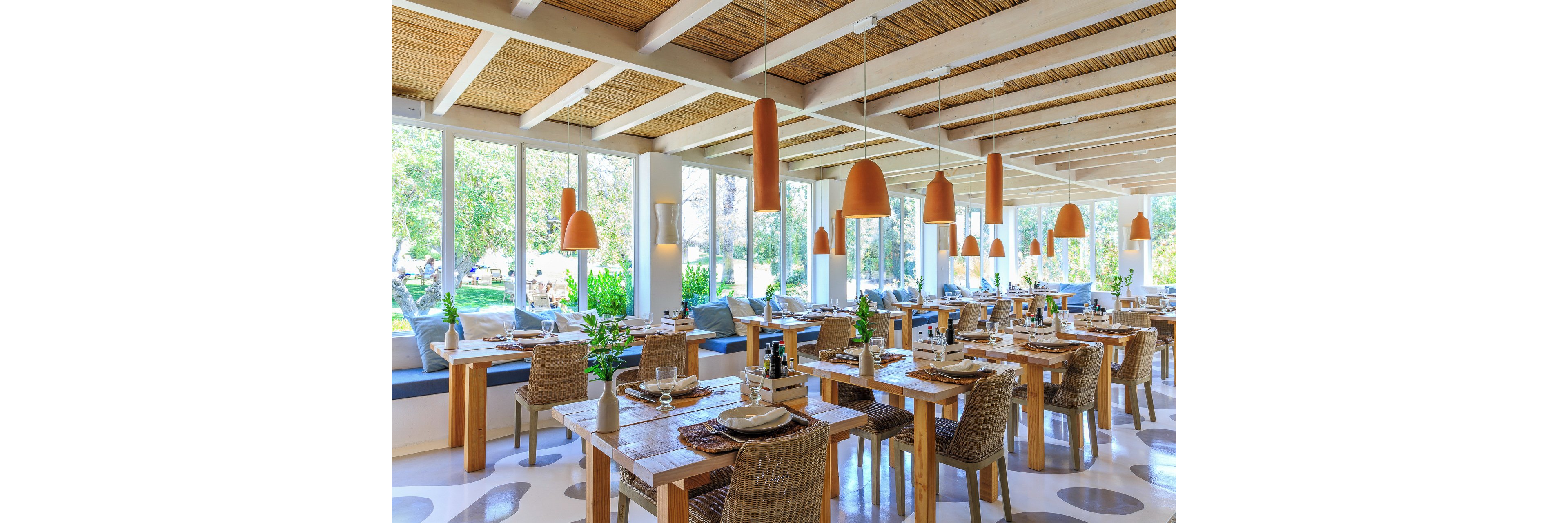 A spacious, modern restaurant dining area features neatly arranged wooden tables and wicker chairs. Large floor-to-ceiling windows let in abundant natural light and reveal lush greenery outside. Warm terracotta pendant lights hang from the ceiling, complementing the natural wood and white color palette. Table settings with plates, glasses, and small plants create an inviting, relaxed atmosphere.