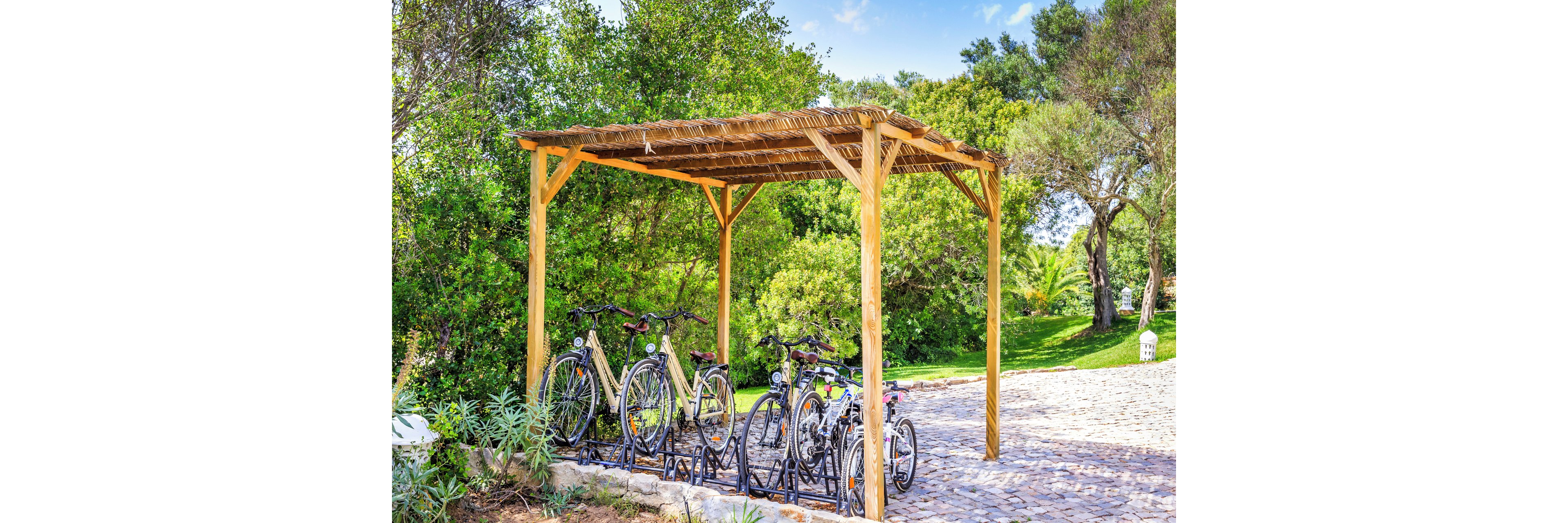 Several bicycles are parked in a metal rack beneath a simple wooden shelter. The structure stands beside a cobblestone path surrounded by lush green trees and shrubs. Bright daylight and clear skies create a vibrant, summery atmosphere. The scene suggests a peaceful park or garden area designed for cyclists.