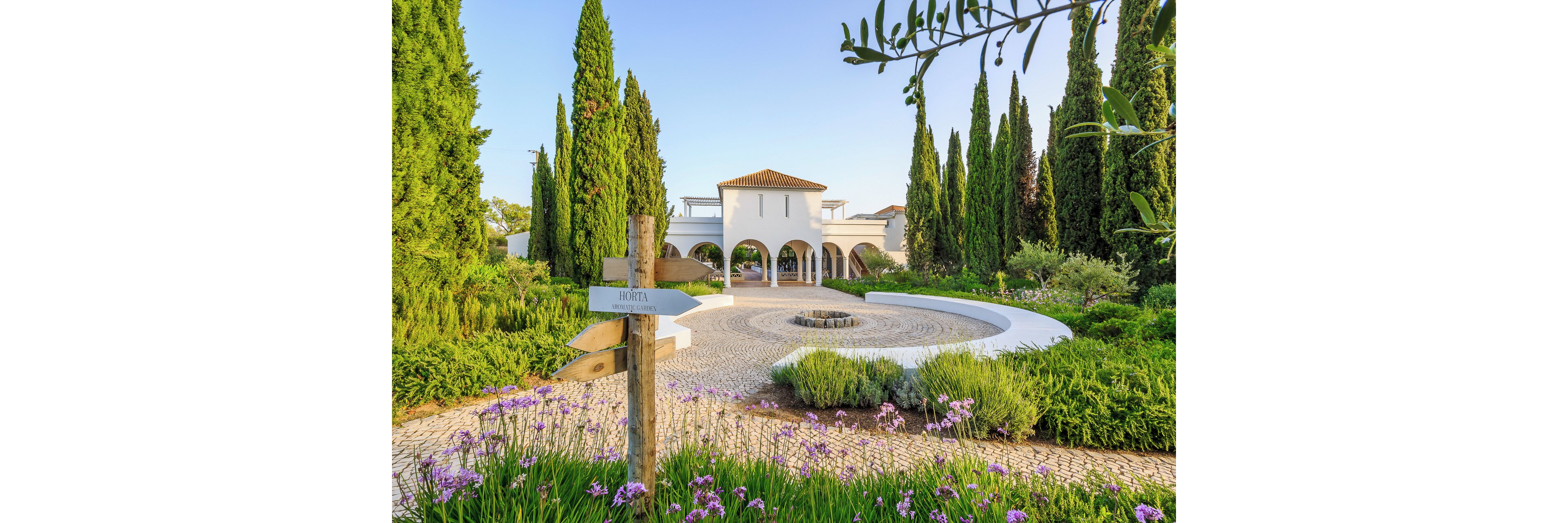 A white Mediterranean-style villa with arched entrances stands at the end of a circular cobblestone driveway. The foreground features a wooden direction sign pointing toward the hotel, surrounded by blooming purple flowers and manicured shrubs. Tall cypress trees frame the scene, creating a serene and elegant atmosphere. Warm sunlight enhances the greenery and architectural details, suggesting a peaceful resort or countryside retreat.