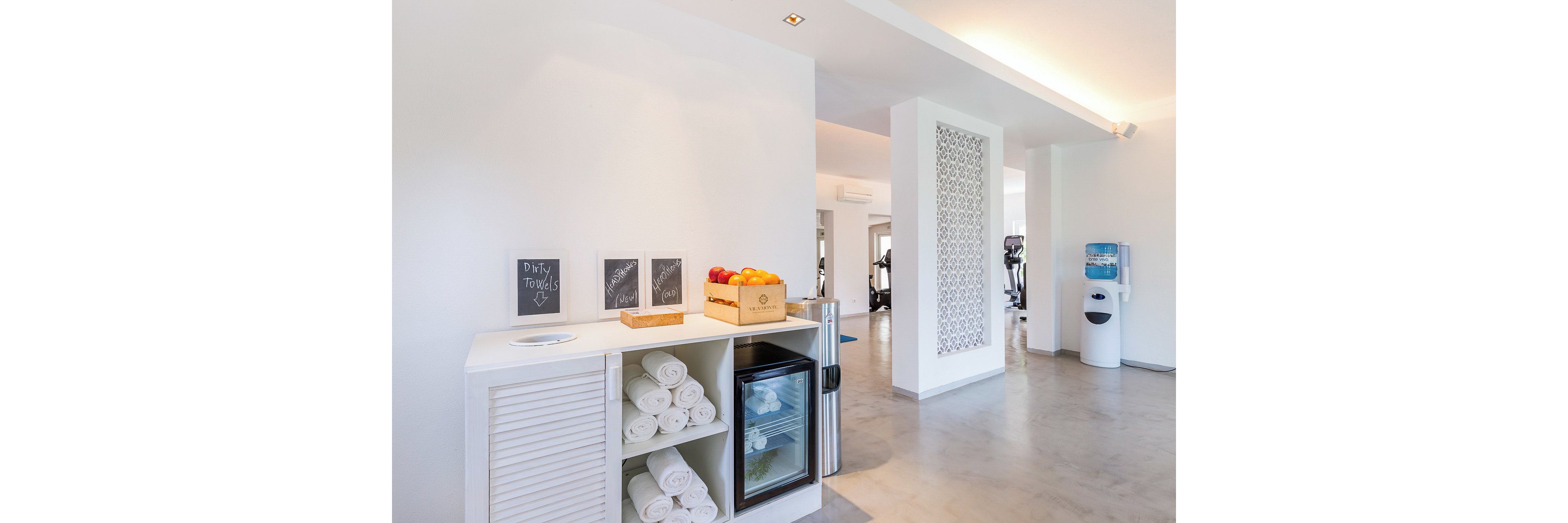 A bright, modern gym refreshment station features neatly rolled white towels stacked beside a small glass-front refrigerator. A wooden crate of fresh apples and chalkboard signs sit on the countertop, adding a welcoming touch. In the background, exercise equipment and a water cooler are visible in the open, minimalist space. Clean white walls, polished floors, and soft lighting create a calm, contemporary atmosphere.