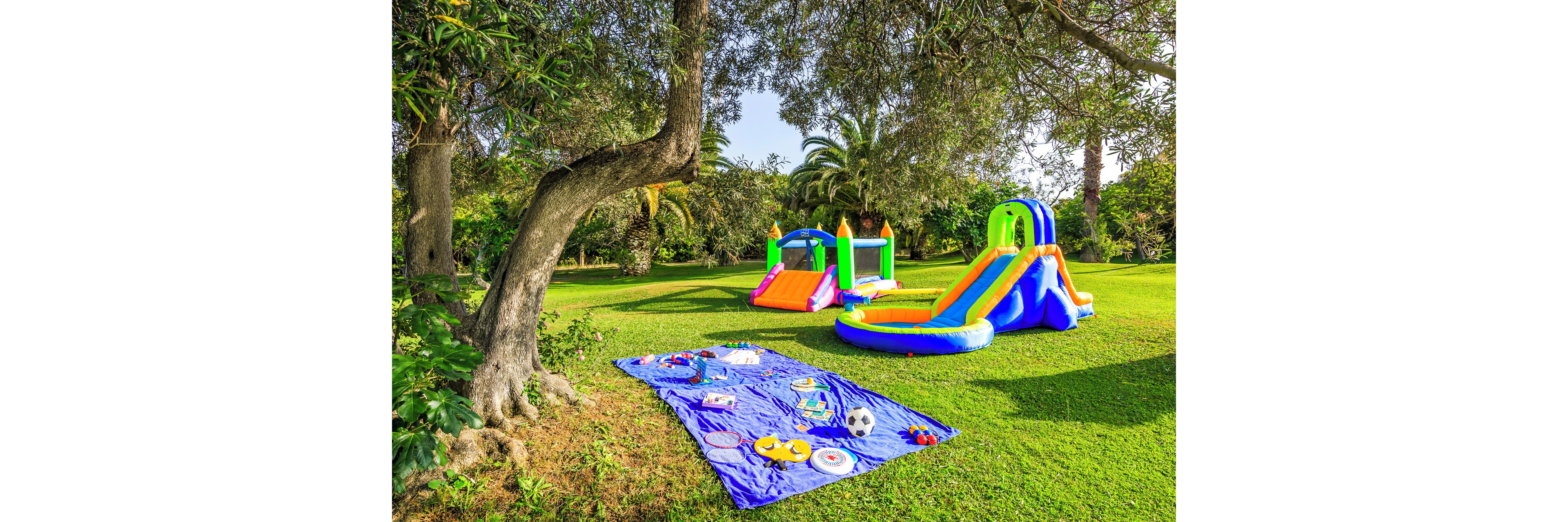A vibrant inflatable playground with slides and climbing sections stands on a lush green lawn. Nearby, a blue picnic blanket is spread out under the shade of a large tree, displaying toys, plates, and outdoor games. Tall palm trees and dense greenery frame the scene, suggesting a warm, sunny day in a spacious backyard or park. The bright primary colors of the inflatables and blanket create a cheerful, family-friendly atmosphere.