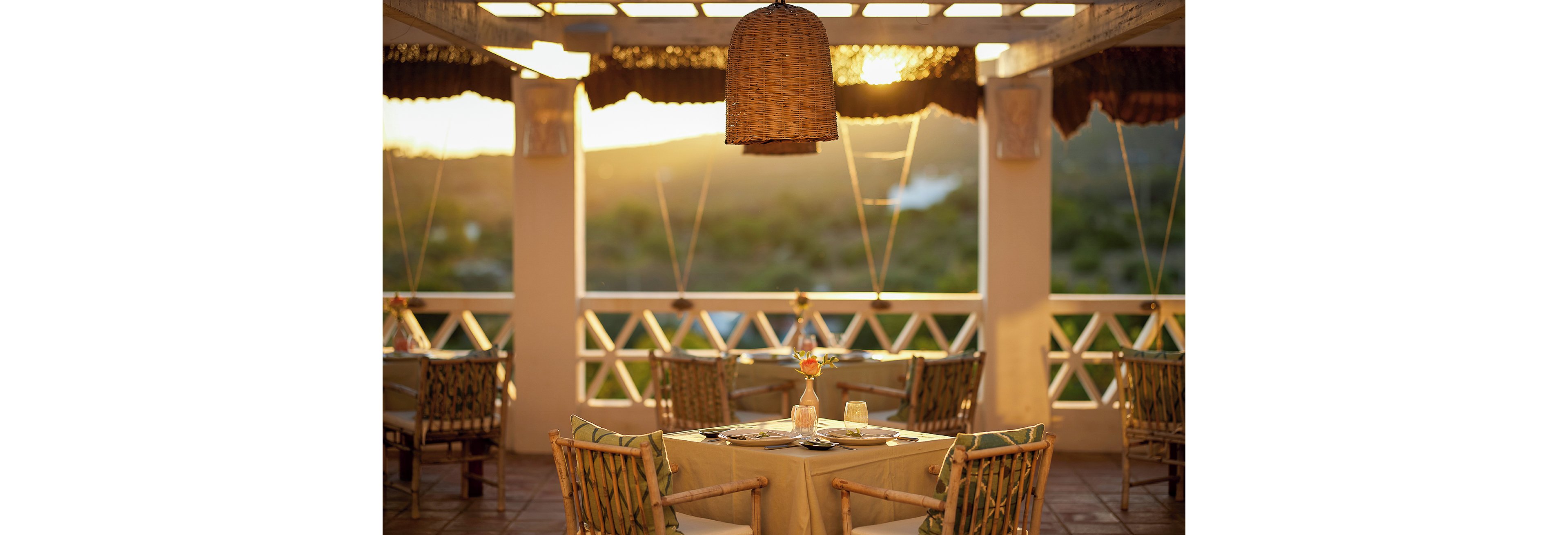 An intimate outdoor restaurant table is set for two on a covered terrace at sunset. Wicker chairs and a central hanging woven lamp create a warm, rustic atmosphere. Soft golden light illuminates the tableware, glassware, and a single flower centerpiece. The open railing reveals a blurred view of greenery and distant hills, enhancing the tranquil, vacation-like setting.