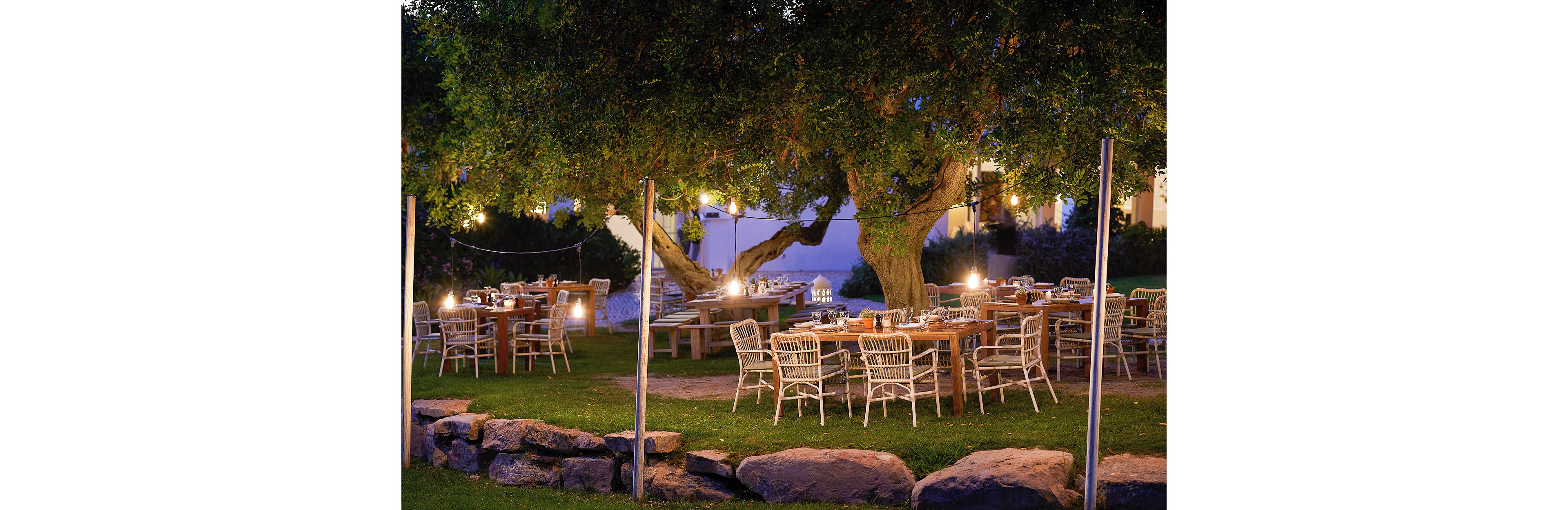 An inviting outdoor dining area is set beneath large trees in a garden at dusk. Wooden tables and white wicker chairs are arranged neatly, ready for guests. Warm string lights and lanterns create a cozy, intimate atmosphere. Stone borders and lush grass frame the scene, suggesting a relaxed, rustic restaurant setting.