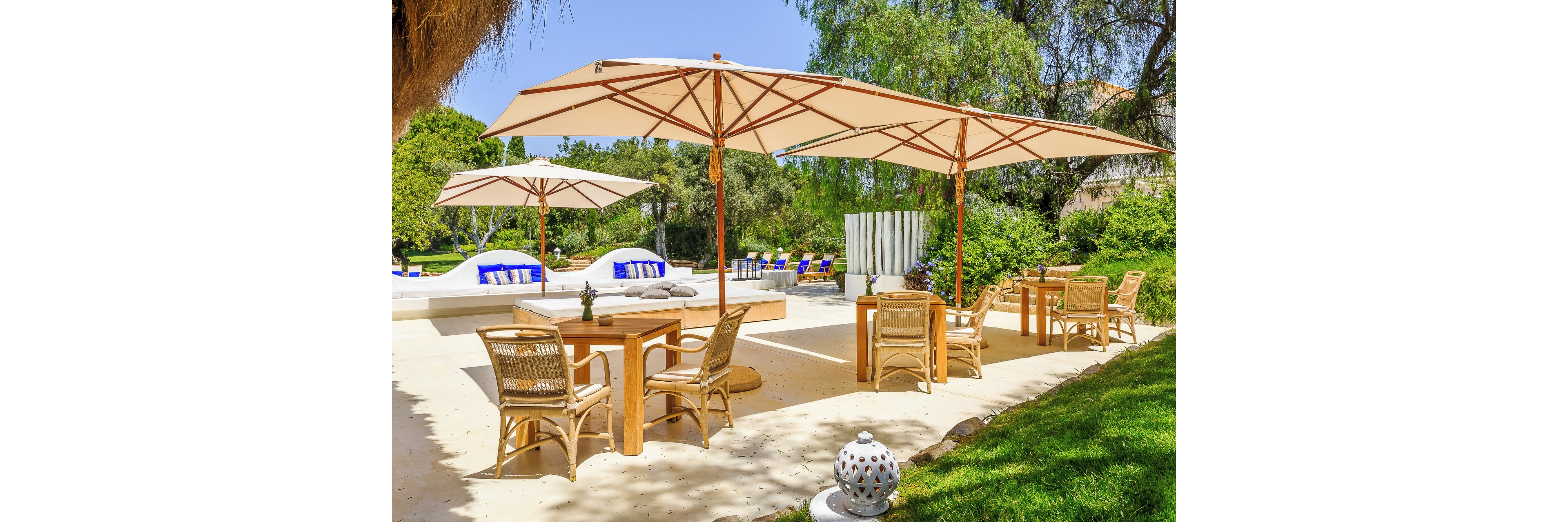 An inviting outdoor patio features wooden tables and wicker chairs arranged under large white umbrellas. In the background, cushioned poolside loungers with blue accents line a raised deck area. Lush green trees and manicured grass surround the space, creating a tranquil resort-like atmosphere. Bright sunlight and clear shadows emphasize the warm, summery setting.