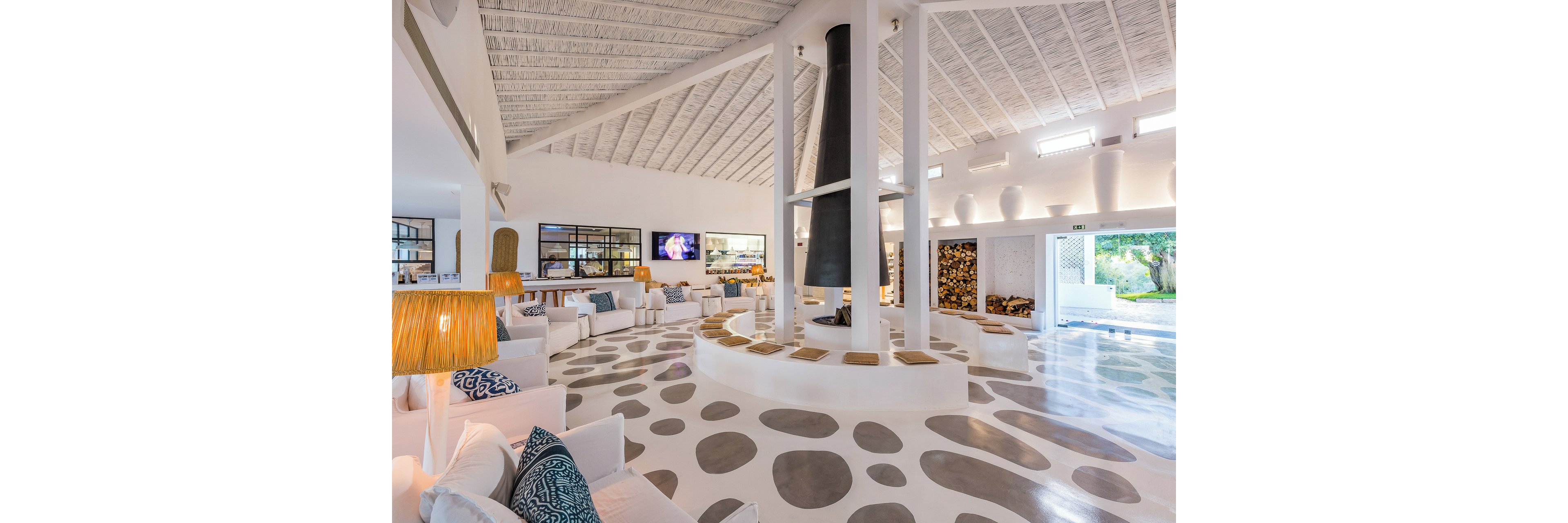 A spacious, contemporary lobby features white walls, high ceilings, and a central circular fireplace with a tall black chimney. Comfortable white sofas with patterned blue cushions are arranged around the seating area, creating an inviting social space. Large windows and glass doors let in abundant natural light, while warm lamps and decorative shelving add visual interest. The polished floor with circular patterns enhances the airy, modern design of the interior.