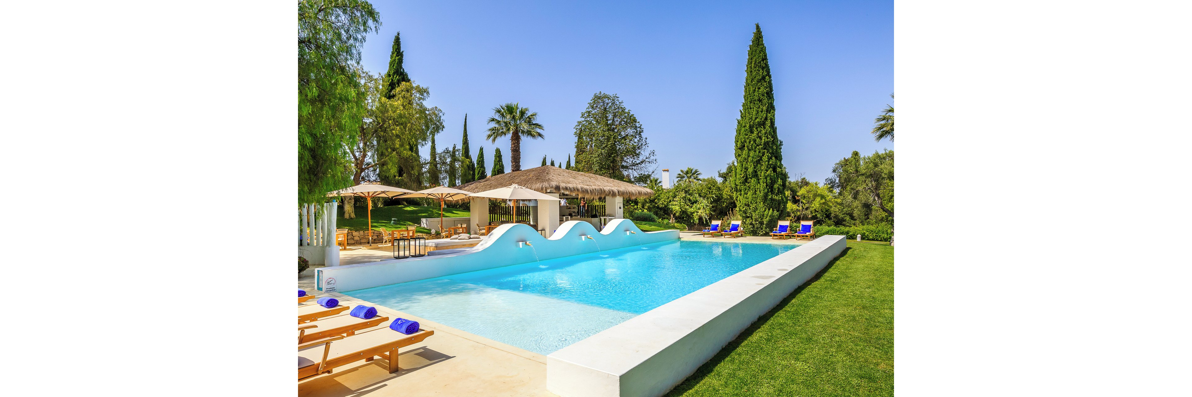 A bright outdoor swimming pool sits in a lush garden setting under a clear blue sky. Wooden loungers with blue towels line the poolside, facing the calm turquoise water. In the background, a shaded bar or lounge area with umbrellas provides a relaxing retreat. Tall palm trees and cypress trees frame the scene, creating a tranquil vacation atmosphere.
