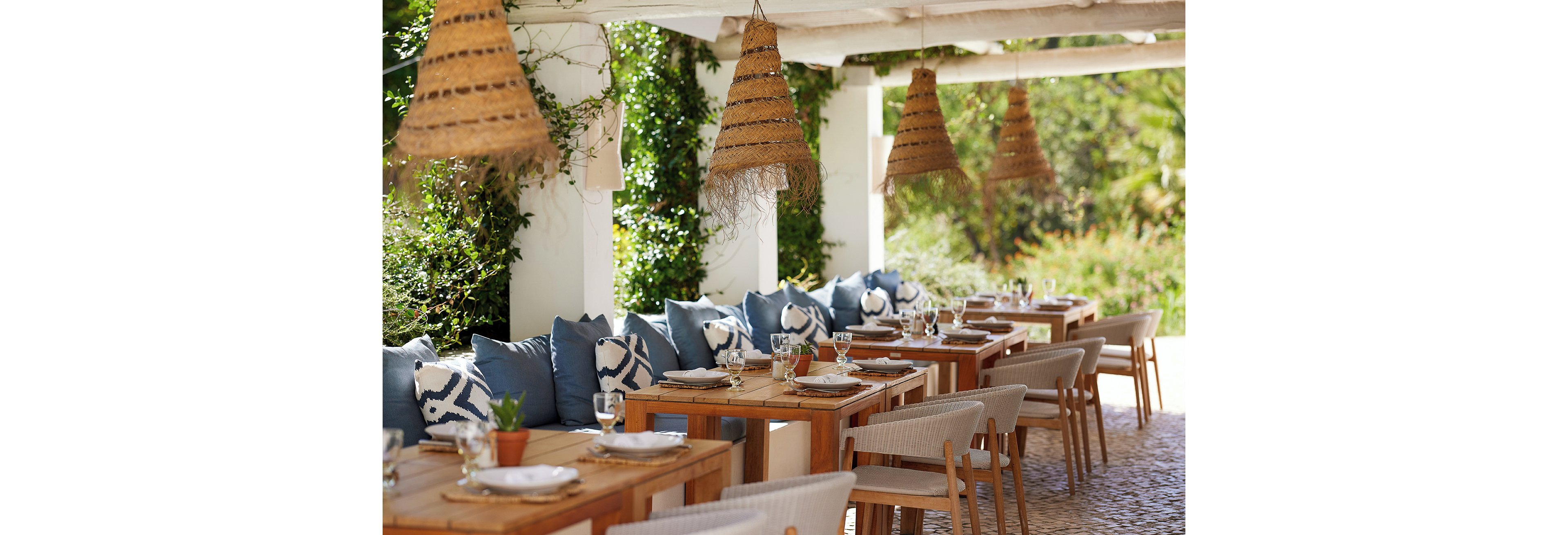 An inviting outdoor restaurant terrace features a long row of wooden tables set for dining. Neutral-toned chairs and blue patterned cushions create a relaxed yet stylish seating area. Woven pendant lights hang from the pergola above, casting a warm, rustic feel. Lush greenery and sunlight in the background enhance the tranquil, Mediterranean-inspired atmosphere.