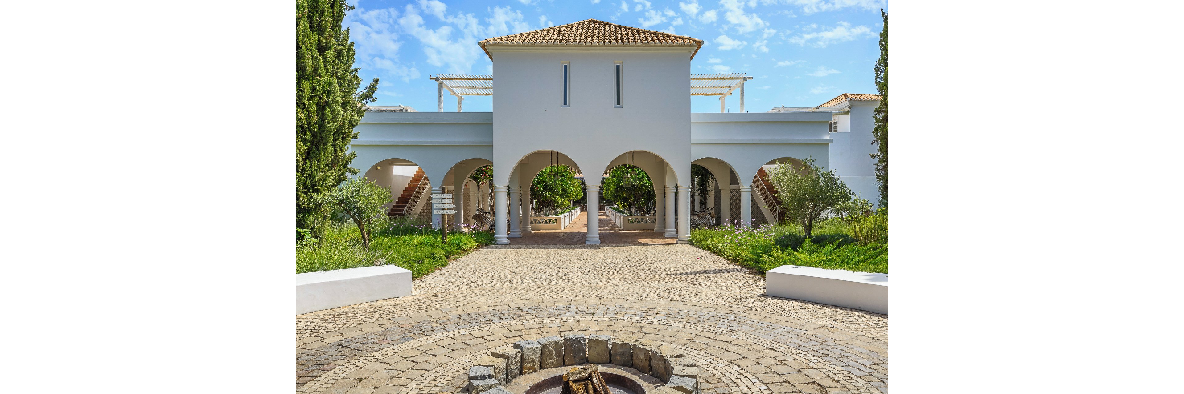 A white Mediterranean-style building with multiple arches forms the focal point of this outdoor courtyard scene. The structure features a tiled roof, symmetrical staircases, and a central walkway leading through the arches. A circular stone fire pit sits in the foreground on a cobblestone surface, surrounded by lush greenery and manicured shrubs. Bright daylight and a blue sky with scattered clouds create a warm, inviting atmosphere.