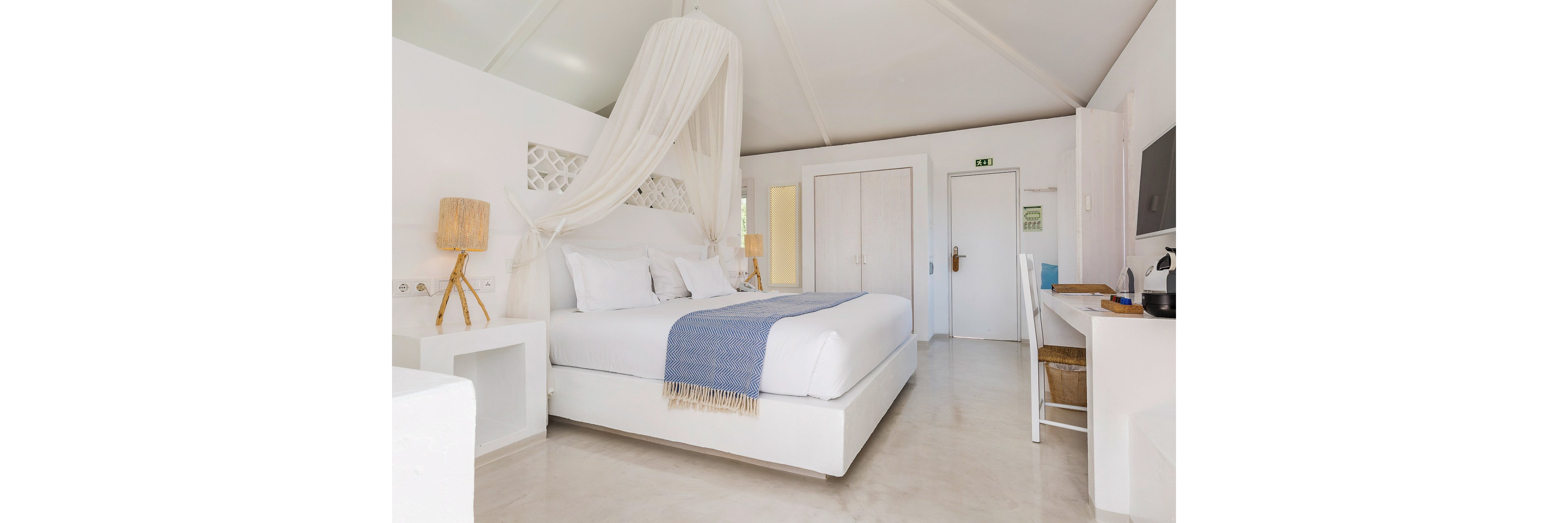 A bright, minimalist bedroom features a white canopy bed with crisp linens and a blue textured throw. The room includes simple bedside tables with woven lamps and a sleek desk area with a chair and television. Neutral walls and flooring enhance the airy, coastal feel of the space. A closed wardrobe and door in the background complete the clean, uncluttered interior.