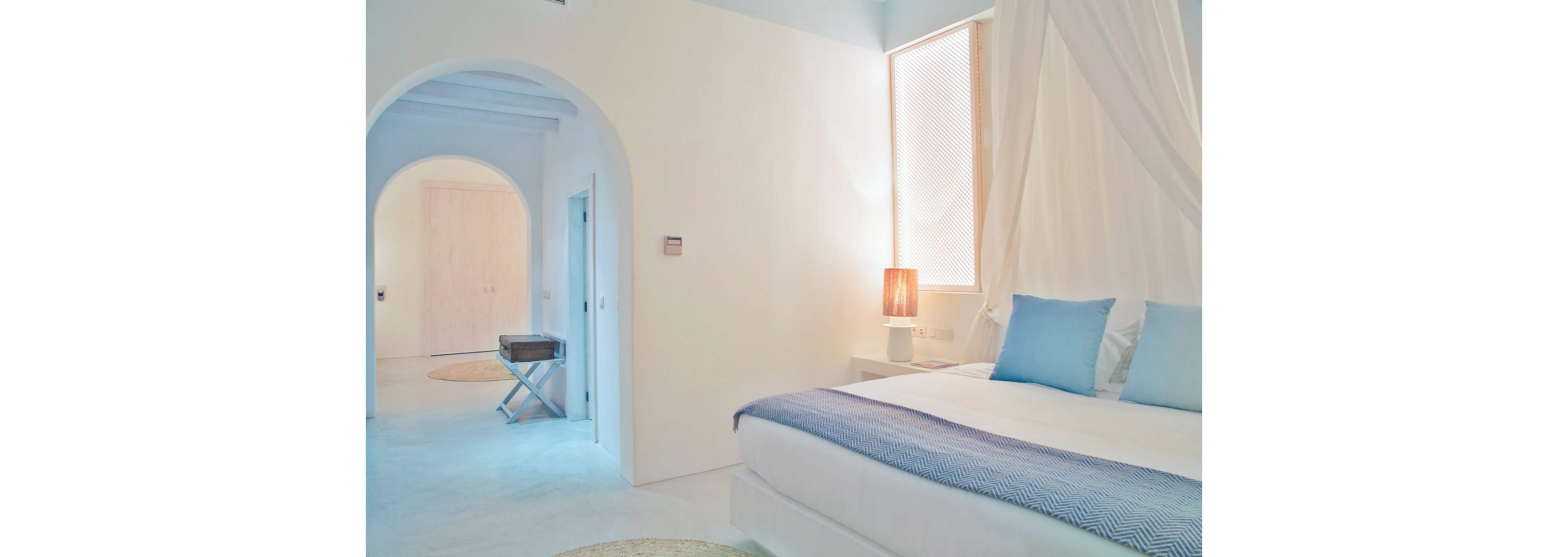 A serene, minimalist bedroom features a neatly made bed with white linens and light blue pillows and throw. Soft natural light filters through a tall window beside a warm glowing table lamp. The room is predominantly white, with smooth walls, a canopy-like drape, and a simple side table. An arched doorway opens to a hallway with a small folding luggage stand, enhancing the airy, spacious feel.