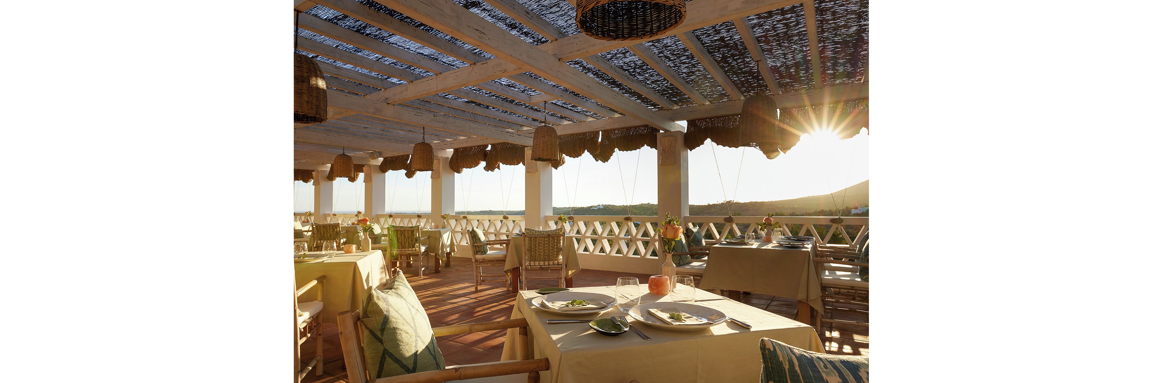 An open-air restaurant terrace is set for dining under a partially shaded pergola roof. Tables are neatly arranged with plates, cutlery, and simple green garnishes, suggesting a relaxed meal service. Warm sunlight streams in from the horizon, casting long shadows and creating a golden glow across the space. Woven pendant lights, wooden furniture, and decorative cushions contribute to a cozy, rustic atmosphere overlooking distant hills.