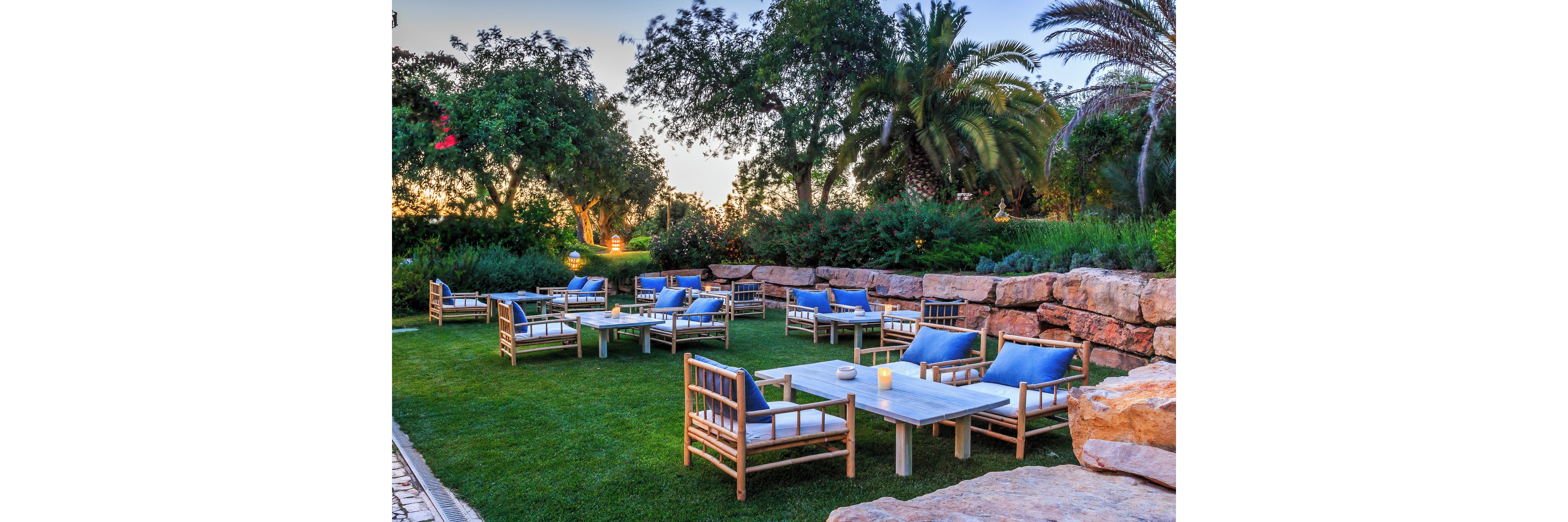 An inviting outdoor lounge area is arranged on a manicured lawn surrounded by lush greenery and palm trees. Wooden chairs with blue cushions are grouped around low stone or concrete tables, creating intimate seating clusters. A stone wall borders the space, adding texture and structure to the garden setting. Soft evening light and small candles on the tables contribute to a relaxed, tranquil atmosphere.