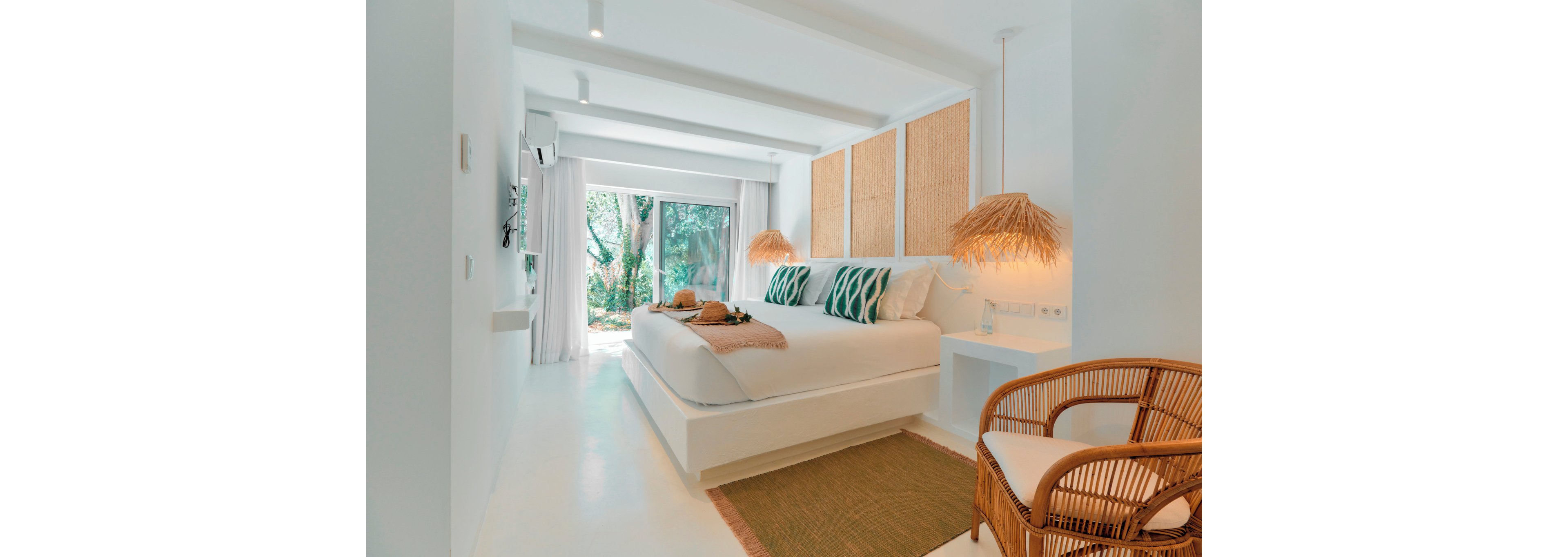 A bright, modern bedroom features a large white bed with patterned green pillows and a soft throw blanket. Natural woven elements, including wall panels, bedside lamps, and a rattan chair, create a relaxed tropical feel. Floor-to-ceiling windows open to a lush garden view, filling the room with daylight. The minimalist white walls and ceiling beams enhance the airy, serene atmosphere.