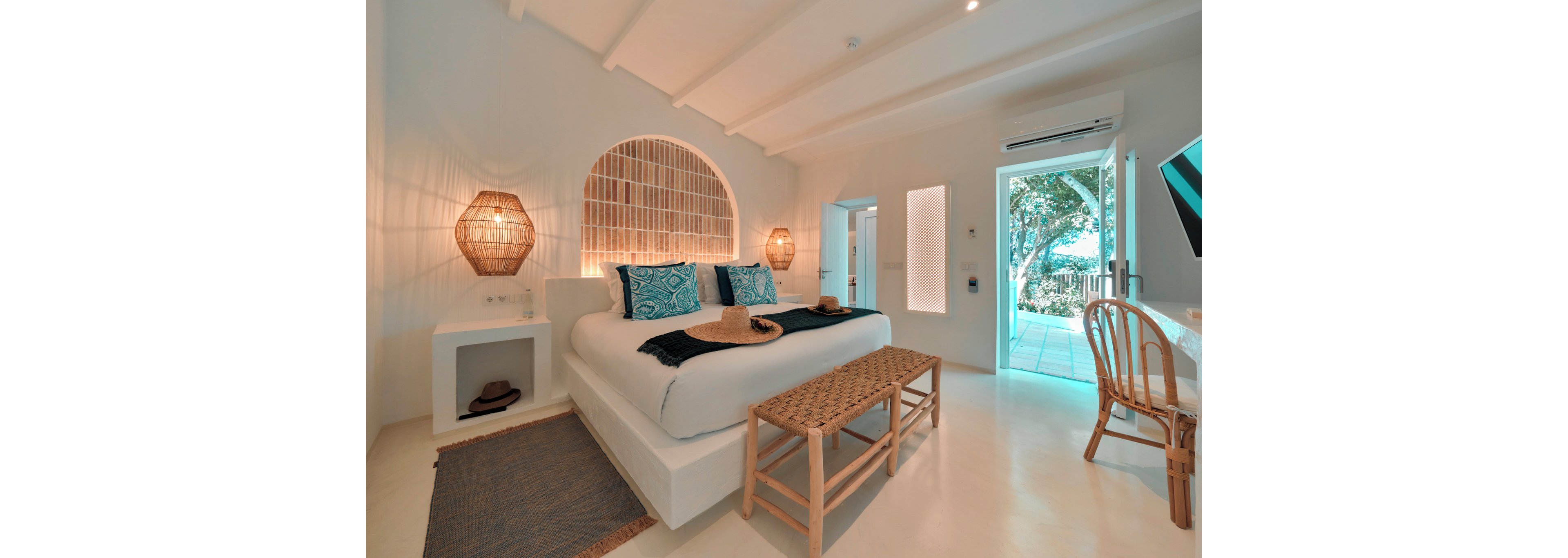 A stylish coastal bedroom features a large white bed with blue patterned pillows and woven hats placed on top. Natural rattan elements, including bedside lamps and a bench at the foot of the bed, add warmth to the bright white interior. A wooden chair and desk sit along one wall, while a doorway opens to a sunlit outdoor area with greenery. Soft lighting and neutral tones create a calm, inviting atmosphere ideal for a beachside retreat.