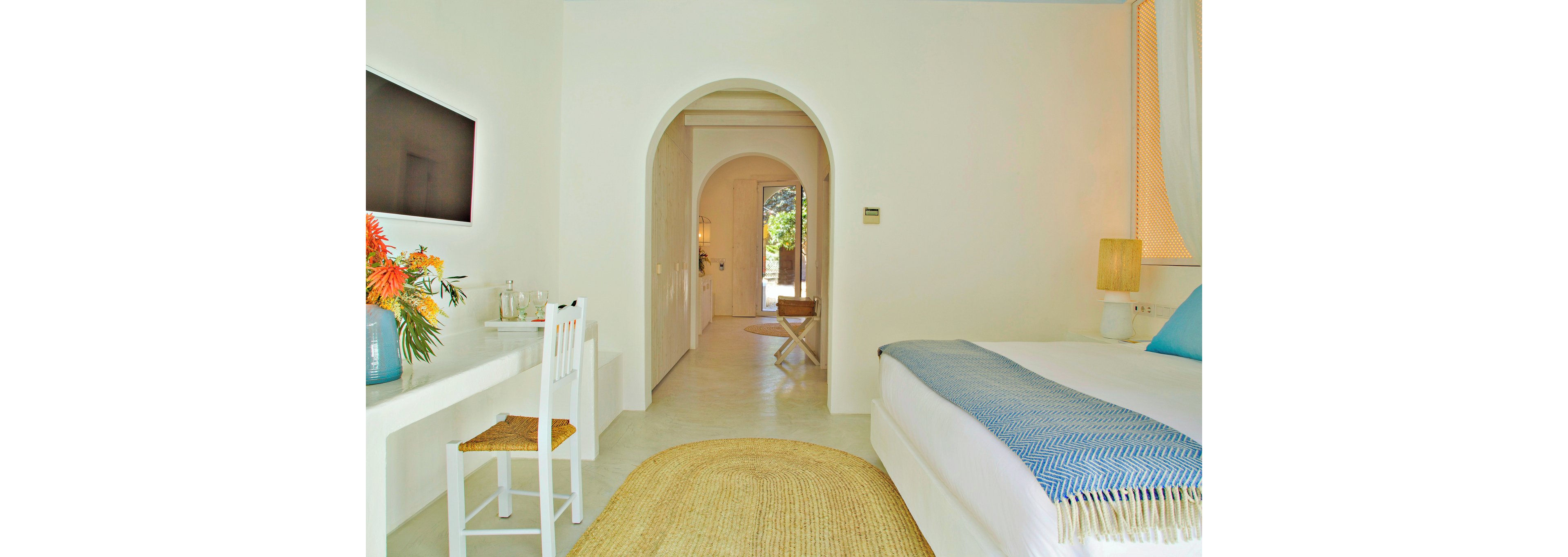 A bright, minimalist bedroom features a neatly made bed with a blue throw and white linens. A woven rug leads toward an arched doorway that opens to a sunlit corridor and outdoor view. On the left, a simple desk with a chair, flowers in a vase, and glassware sits beneath a wall-mounted television. Warm accent lighting and natural textures create a calm, inviting atmosphere.