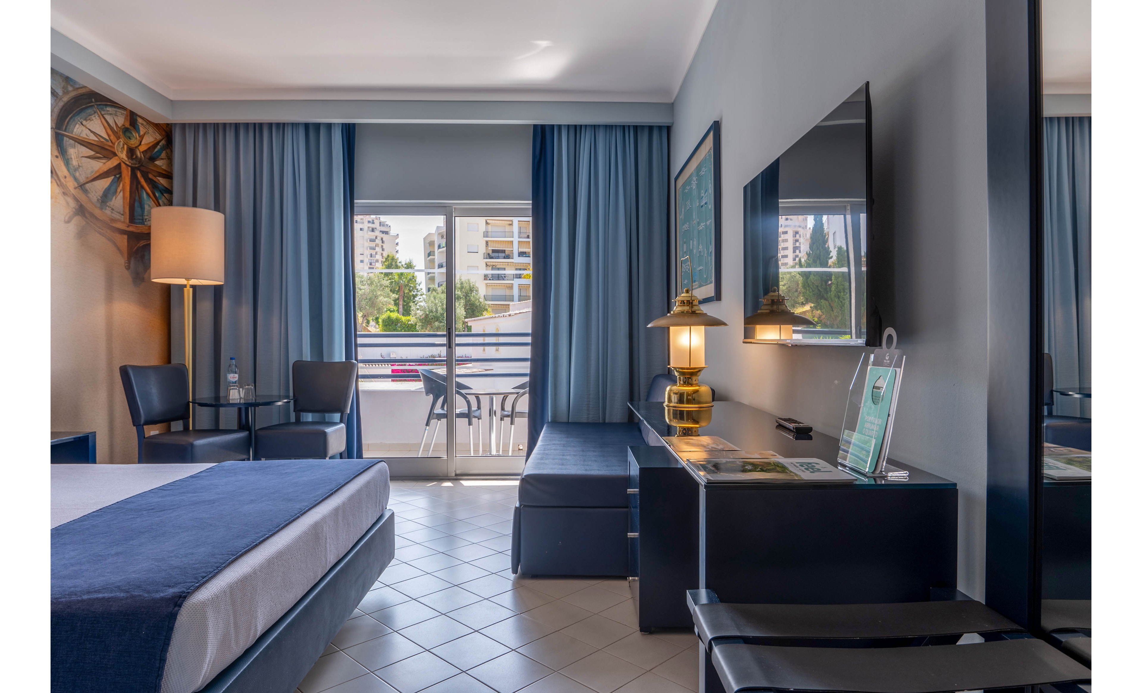 A contemporary hotel room featuring a neatly made bed, a desk with a lamp, and a flat-screen TV. The room opens to a balcony with outdoor seating and a view of urban buildings and greenery. The decor includes blue curtains, a nautical-themed wall piece, and a tiled floor, creating a clean and inviting atmosphere.