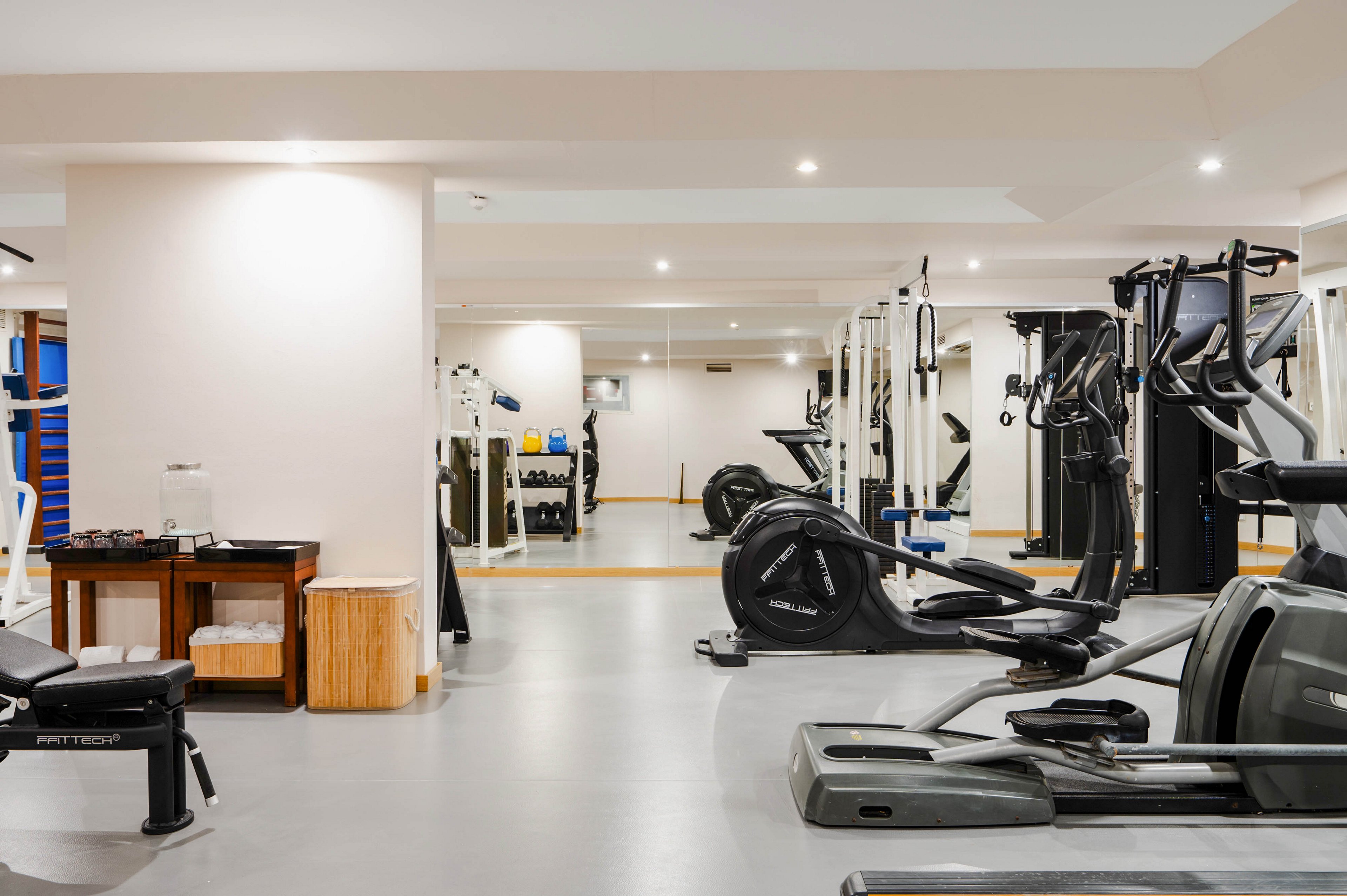 A bright, modern fitness room features various exercise machines and workout stations. Cardio equipment such as treadmills and elliptical trainers are arranged alongside strength-training machines and benches. Large mirrors line the walls, visually expanding the space and reflecting the equipment. The neutral color palette and clean, uncluttered layout create a professional and inviting workout environment.