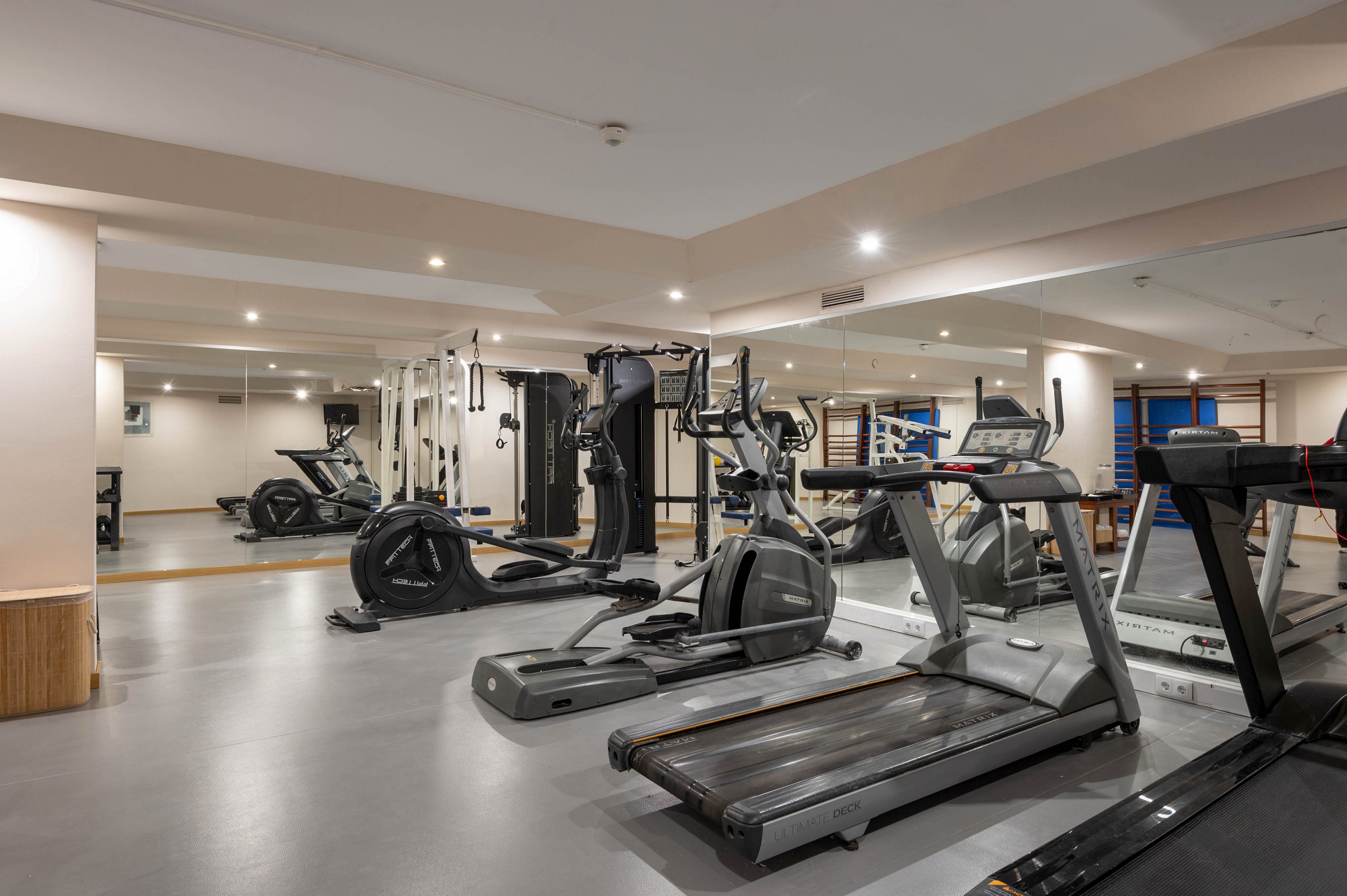 A spacious indoor gym features several treadmills, elliptical machines, and stationary bikes arranged neatly on a smooth gray floor. Large mirrors line the walls, reflecting the exercise equipment and making the room appear bigger and brighter. The ceiling has recessed lighting that creates a clean, contemporary atmosphere. No people are present, emphasizing the equipment and tidy workout space.