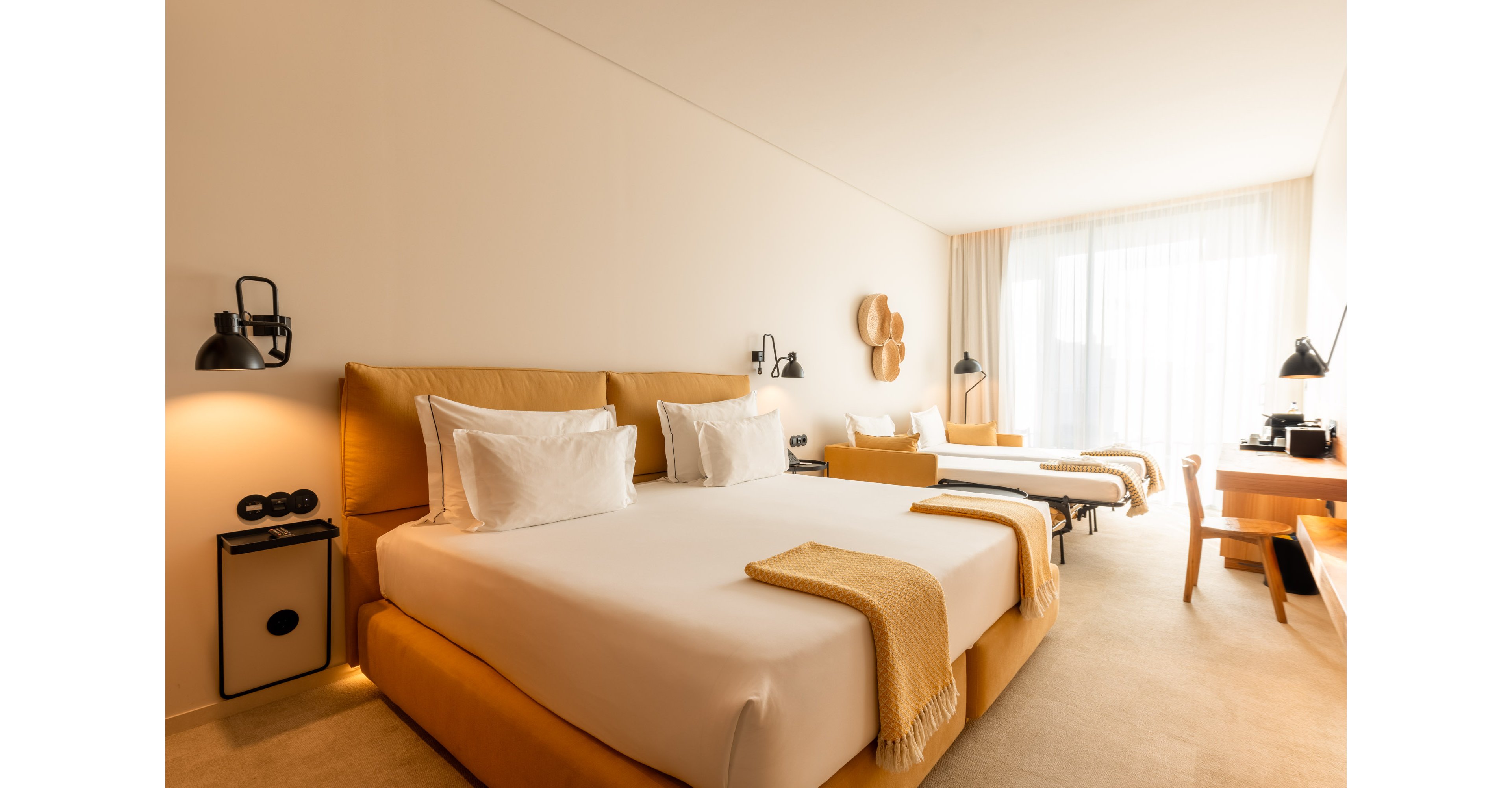 A spacious modern hotel room features a large double bed in the foreground and a single bed in the background. The space is decorated in warm beige and white tones with matching throws and pillows. Large floor-to-ceiling windows with sheer curtains allow natural light to fill the room. Minimalist furniture, bedside lamps, and a work desk create a clean, contemporary atmosphere.