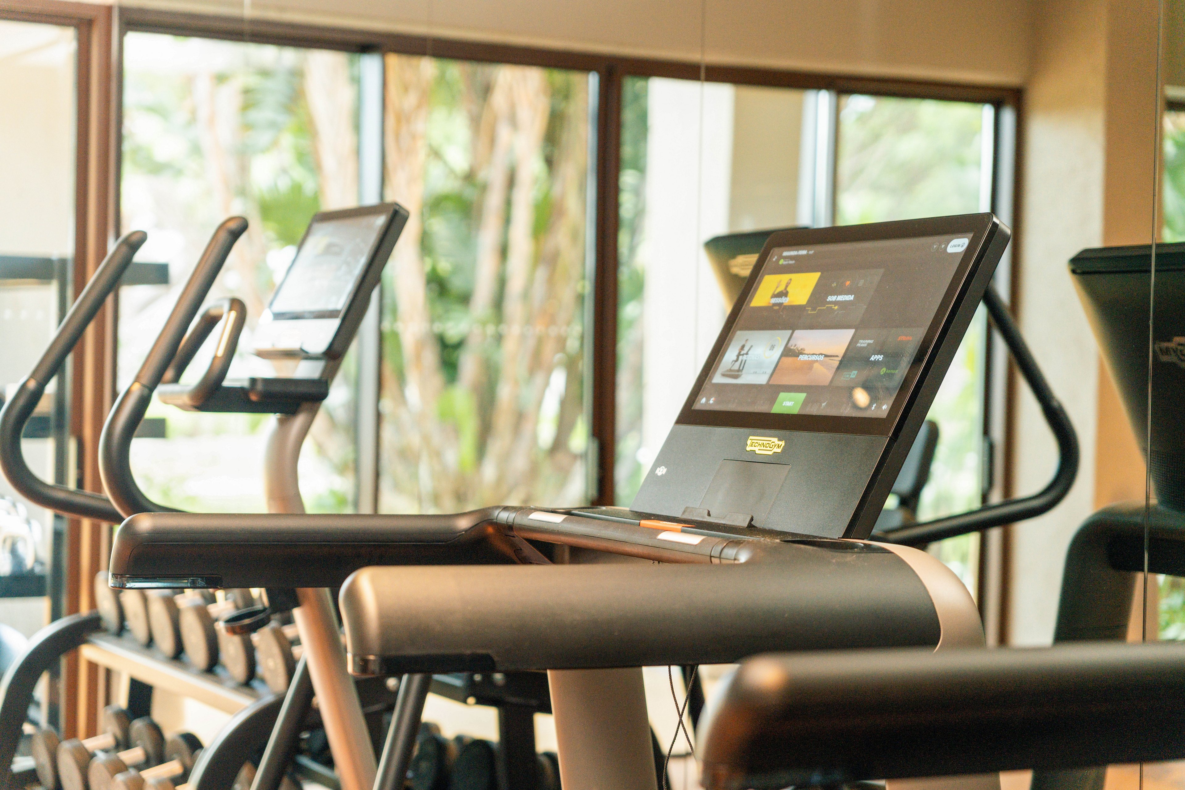 A stationary exercise bike features a large digital touchscreen console in a bright fitness studio. The display shows multiple content tiles and workout options, suggesting interactive training or entertainment. In the background, another cardio machine and a rack of dumbbells are visible, emphasizing a well-equipped gym environment. Natural light streams through large windows, creating a clean and contemporary workout atmosphere.