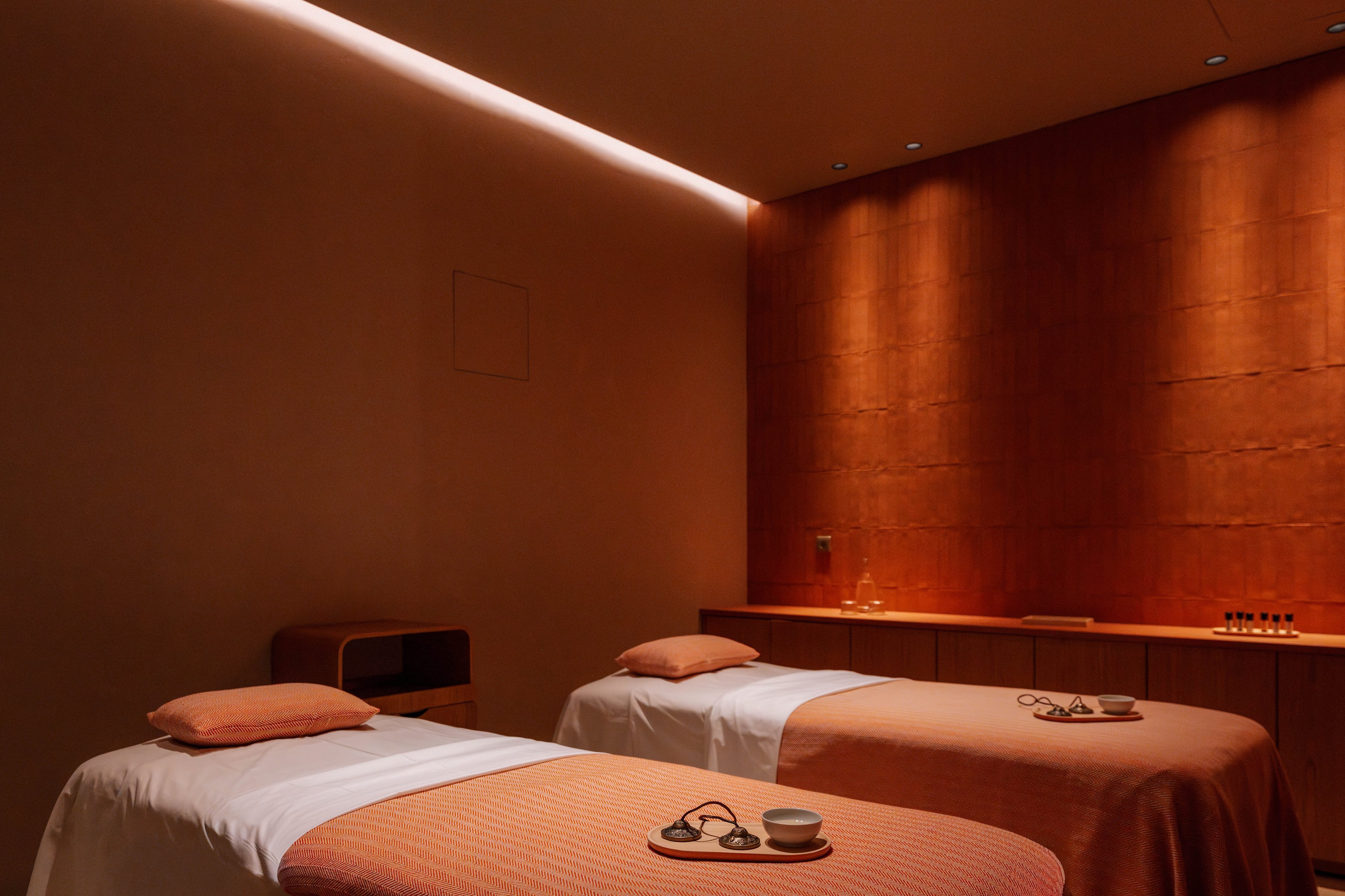 A tranquil spa treatment room features two neatly made massage beds with soft pillows and matching peach-colored covers. The space is warmly lit, highlighting a textured wall and minimalist wooden cabinetry along the side. Small bowls and massage stones rest on the beds, suggesting a relaxing wellness ritual. The overall atmosphere is calm, intimate, and designed for peaceful rejuvenation.