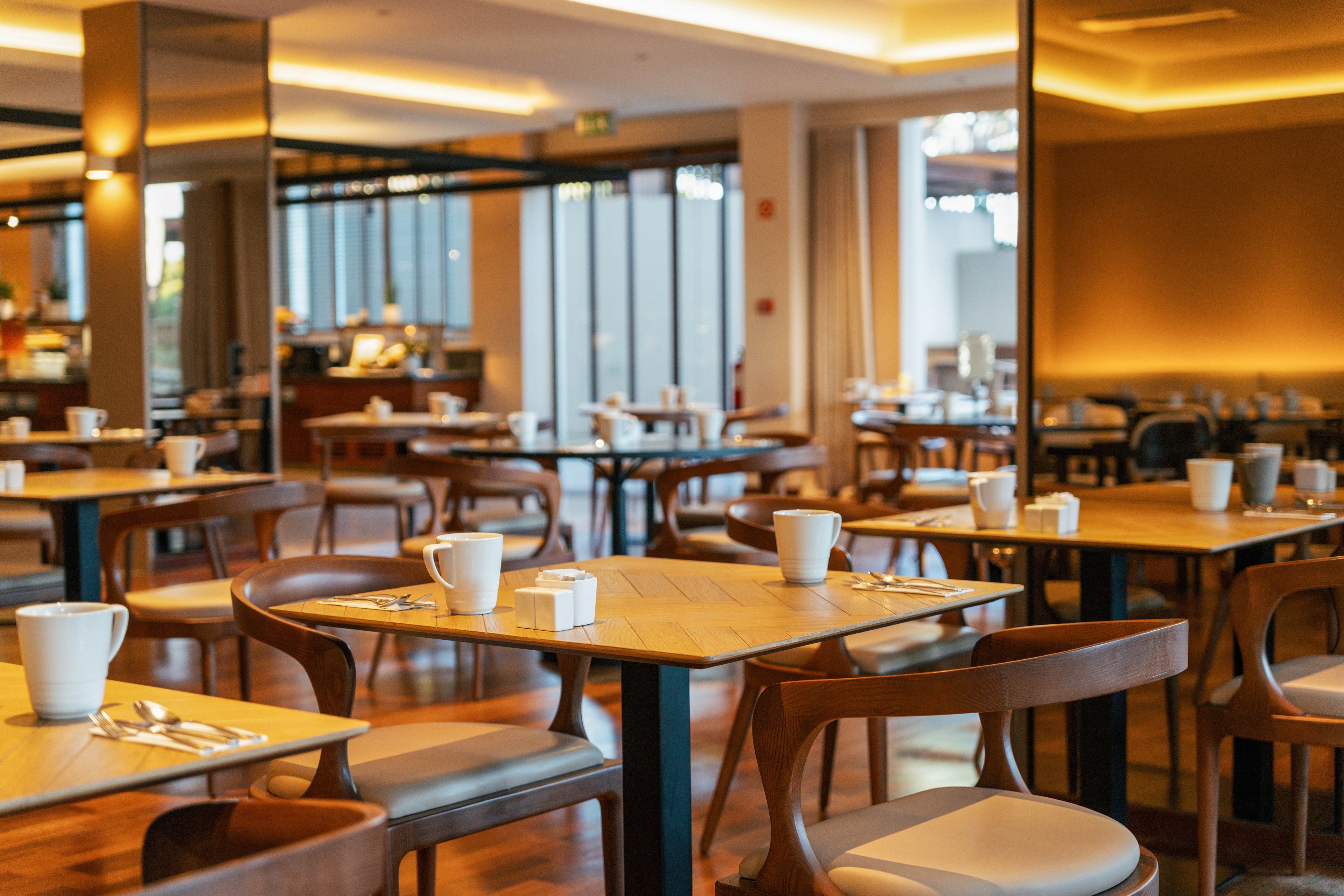 A contemporary restaurant dining area features neatly arranged wooden tables and chairs. White coffee cups and cutlery are set on each table, suggesting preparation for breakfast or a casual meal. Large windows and glass doors allow natural light to fill the space, highlighting the warm tones of the interior. The room appears clean, quiet, and ready to welcome guests.