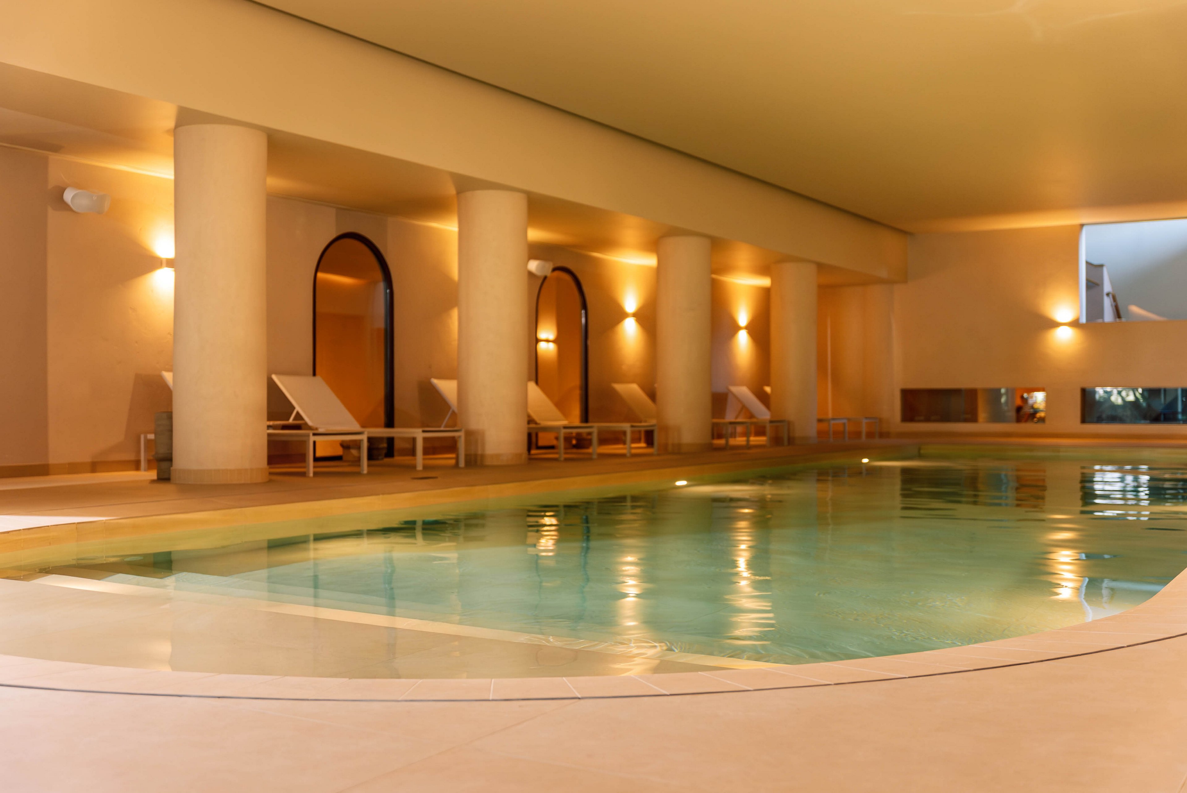 A woman in a black swimsuit relaxes at the edge of a calm indoor swimming pool. The spa-like setting features warm lighting, arched doorways, and white columns. Lounge chairs line the far side of the pool, enhancing the atmosphere of comfort and leisure. The overall scene conveys tranquility and upscale wellness.