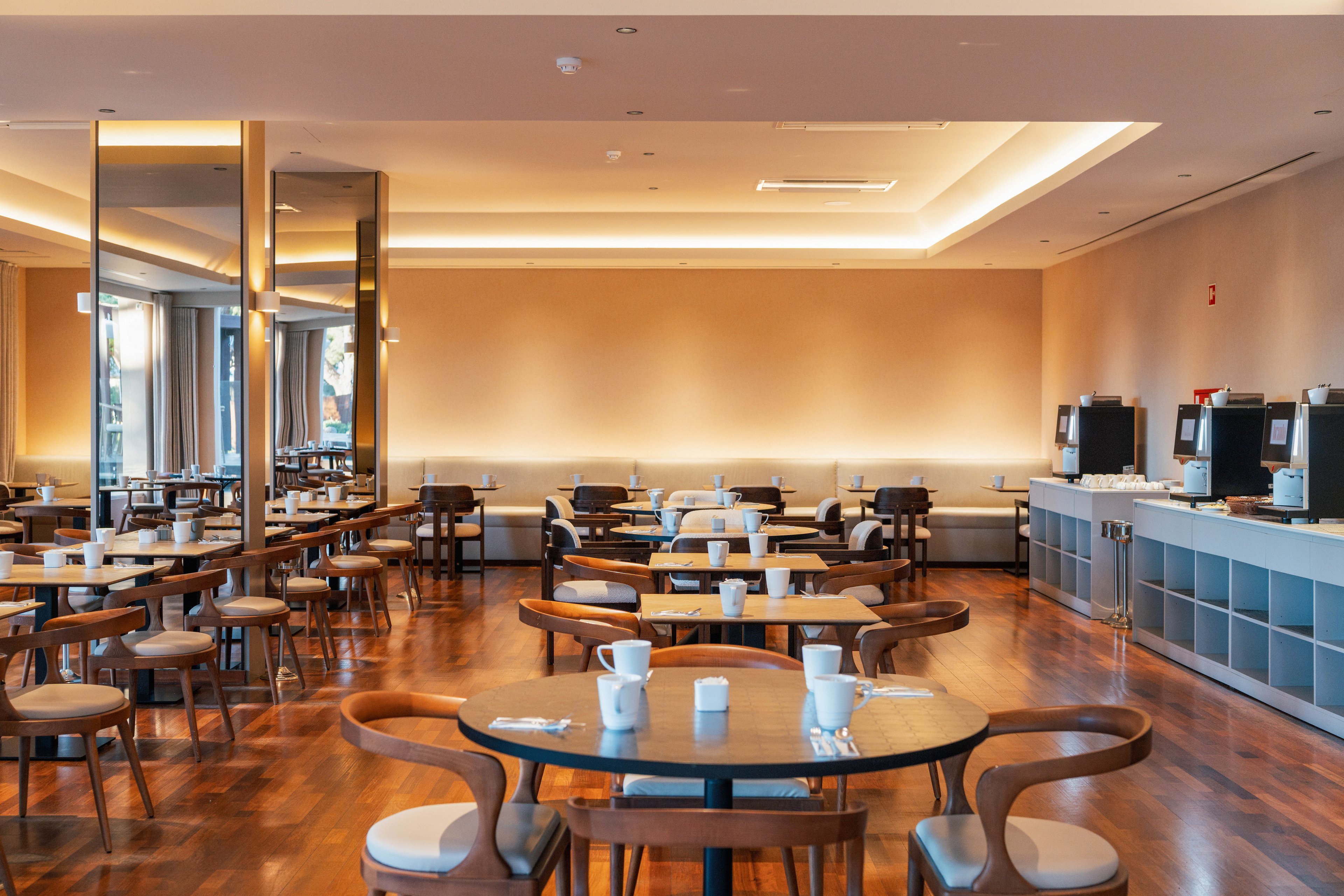 A contemporary dining room features round wooden tables and curved chairs neatly arranged in rows. White cups and saucers are placed on the tables, suggesting preparation for breakfast or coffee service. The space has warm ambient lighting, a polished wooden floor, and large mirrors that visually expand the room. On the right side, a self-service counter with beverage machines and storage cabinets completes the hotel restaurant setting.