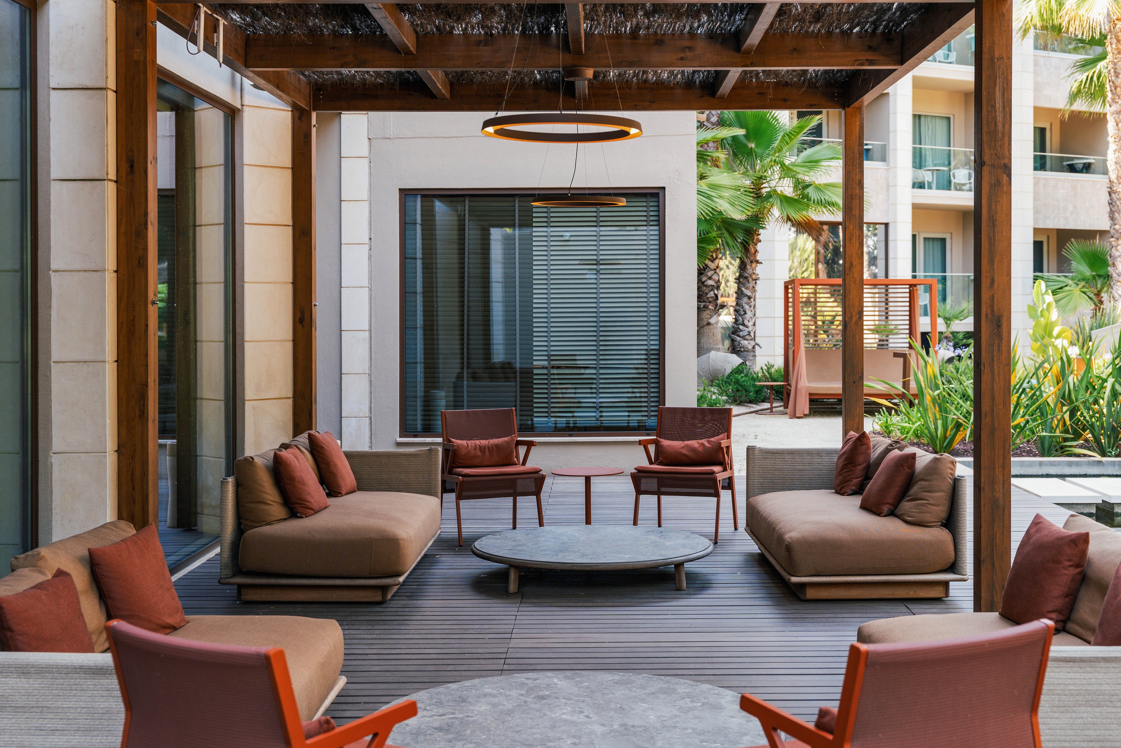 Contemporary outdoor lounge space features cushioned sofas and armchairs arranged around a low circular coffee table. The area is set beneath a wooden pergola, adjacent to large glass doors and windows of a modern building. Warm earth tones dominate the furniture and pillows, complemented by greenery and palm trees in the background. The scene suggests a relaxed, comfortable atmosphere suitable for socializing or quiet retreat.