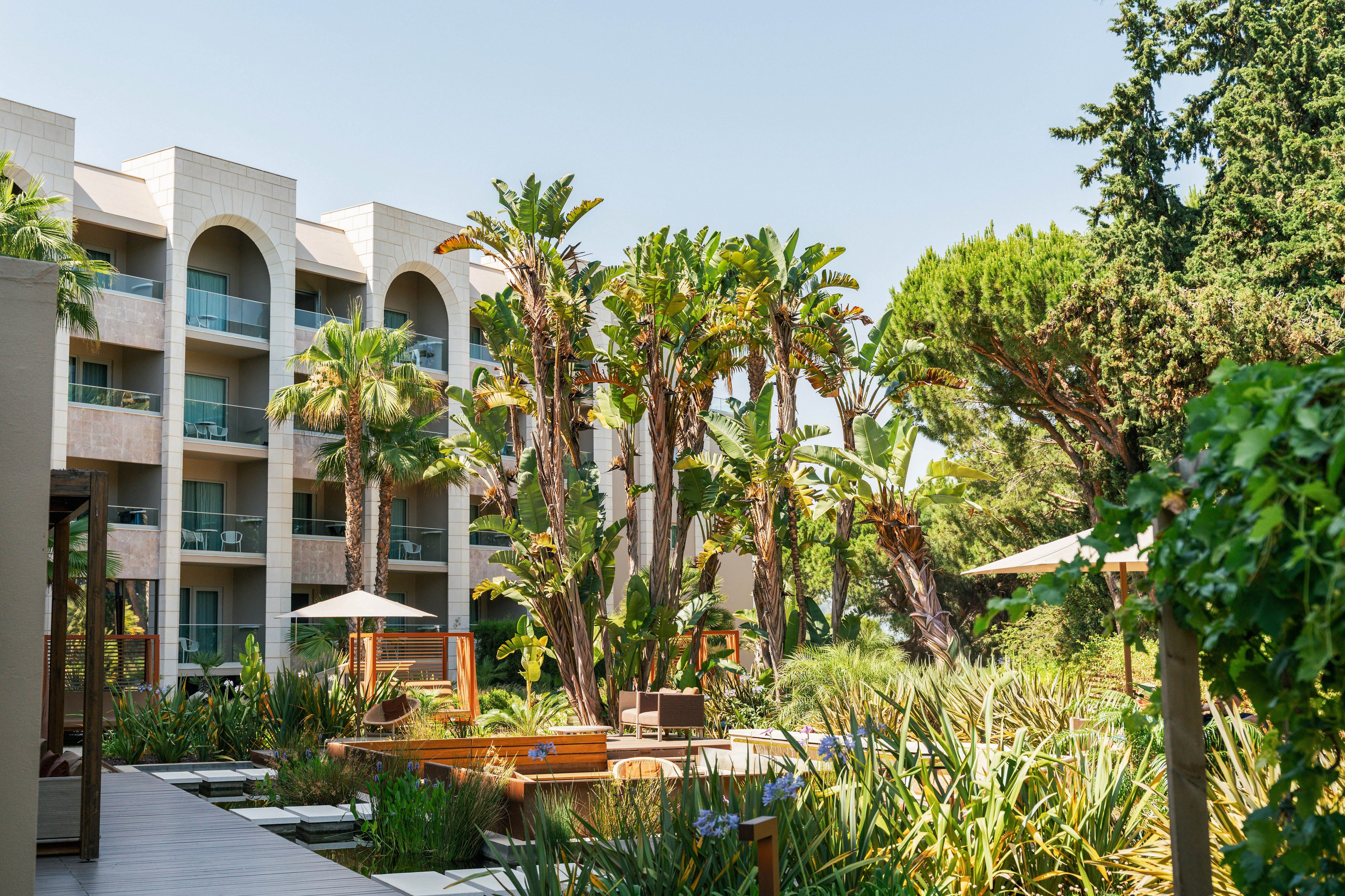 A contemporary multi-story resort building overlooks a lush courtyard filled with tropical plants and tall palm trees. Wooden walkways and seating areas with sun umbrellas create a relaxing outdoor space. The scene is brightly lit under clear skies, emphasizing the greenery and architectural arches. The overall atmosphere suggests a tranquil vacation or leisure setting.
