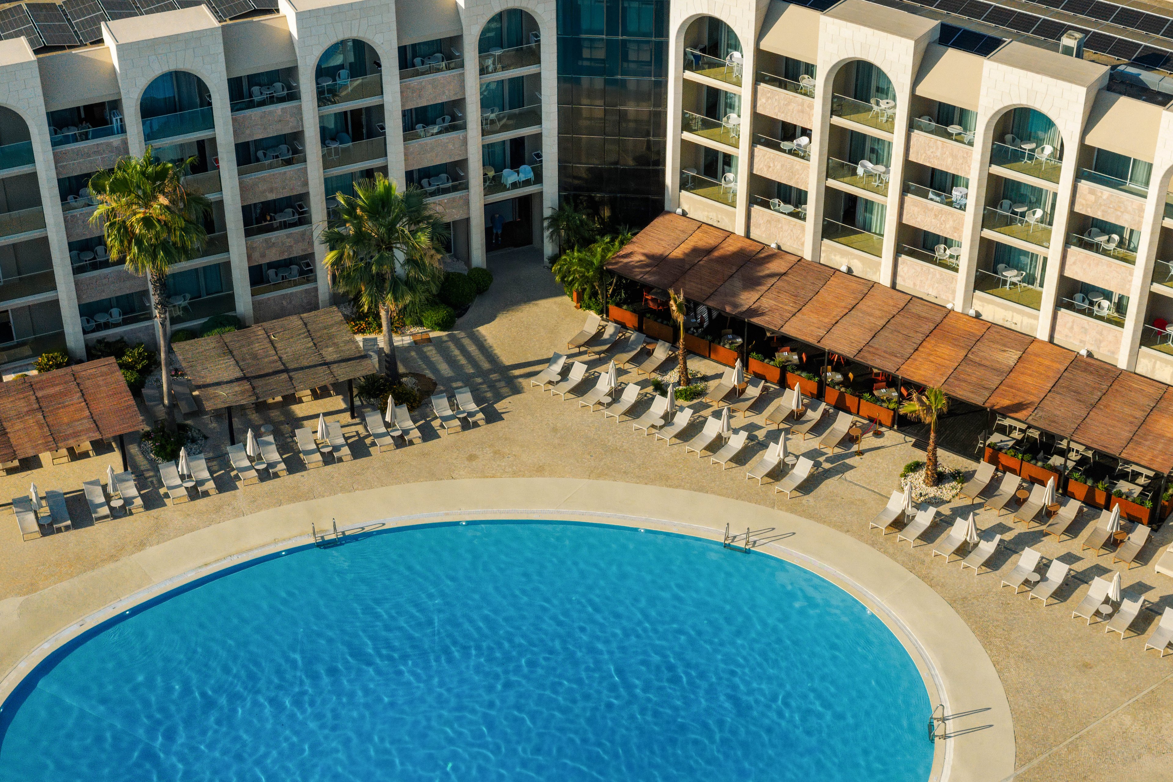 A large curved swimming pool is surrounded by rows of sun loungers at a resort hotel. The multi-story building features balconies overlooking the pool area, with palm trees adding a tropical touch. Wooden pergolas provide shaded seating along one side of the deck. The scene suggests a sunny vacation setting focused on relaxation and leisure.