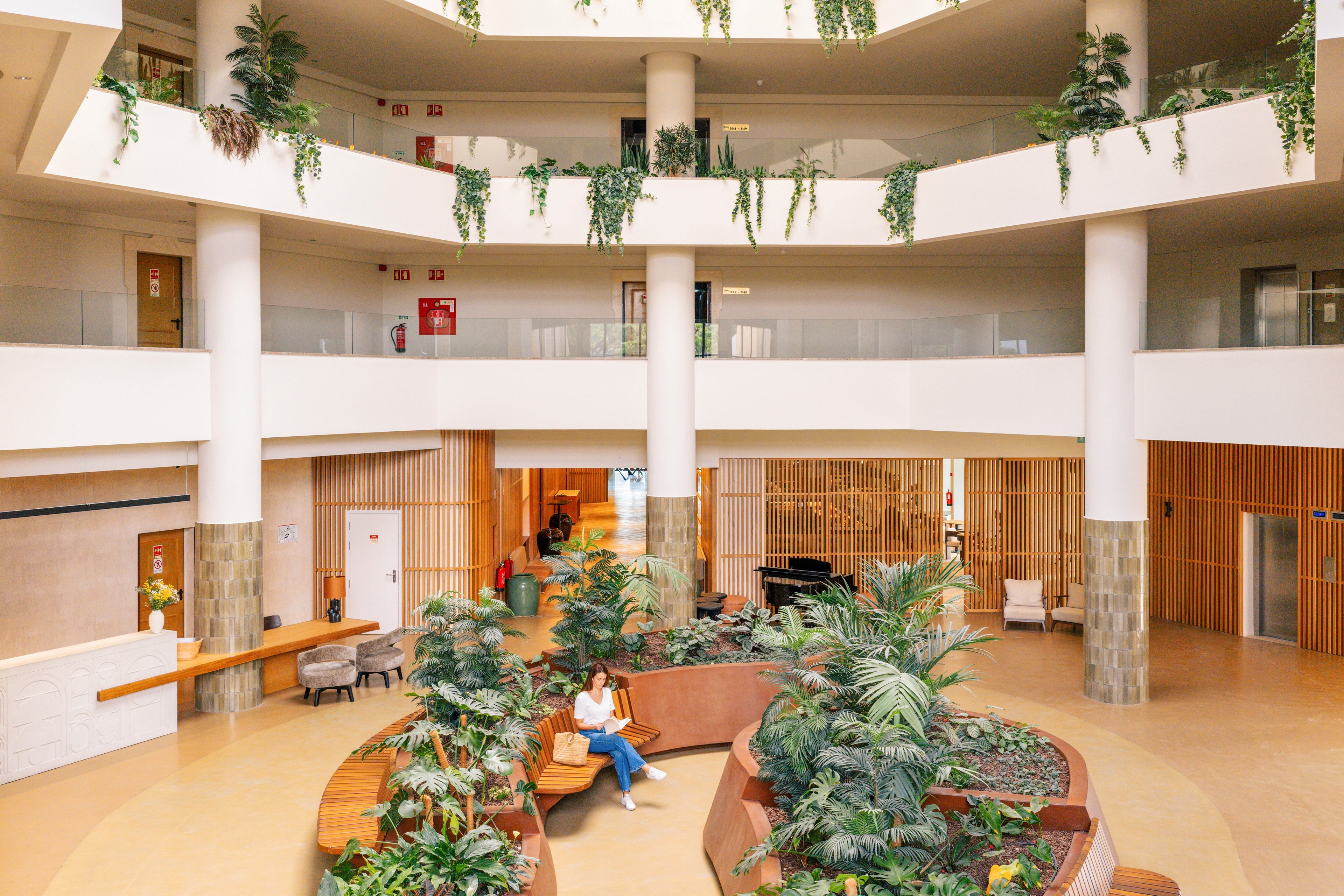 A spacious multi-level atrium features a central seating area surrounded by lush indoor plants. A person sits on a curved bench, reading or using a device in the bright, open lounge. Warm wood accents, light-colored floors, and white columns create a contemporary, welcoming atmosphere. Natural light and hanging greenery enhance the relaxed, biophilic design of the interior space.