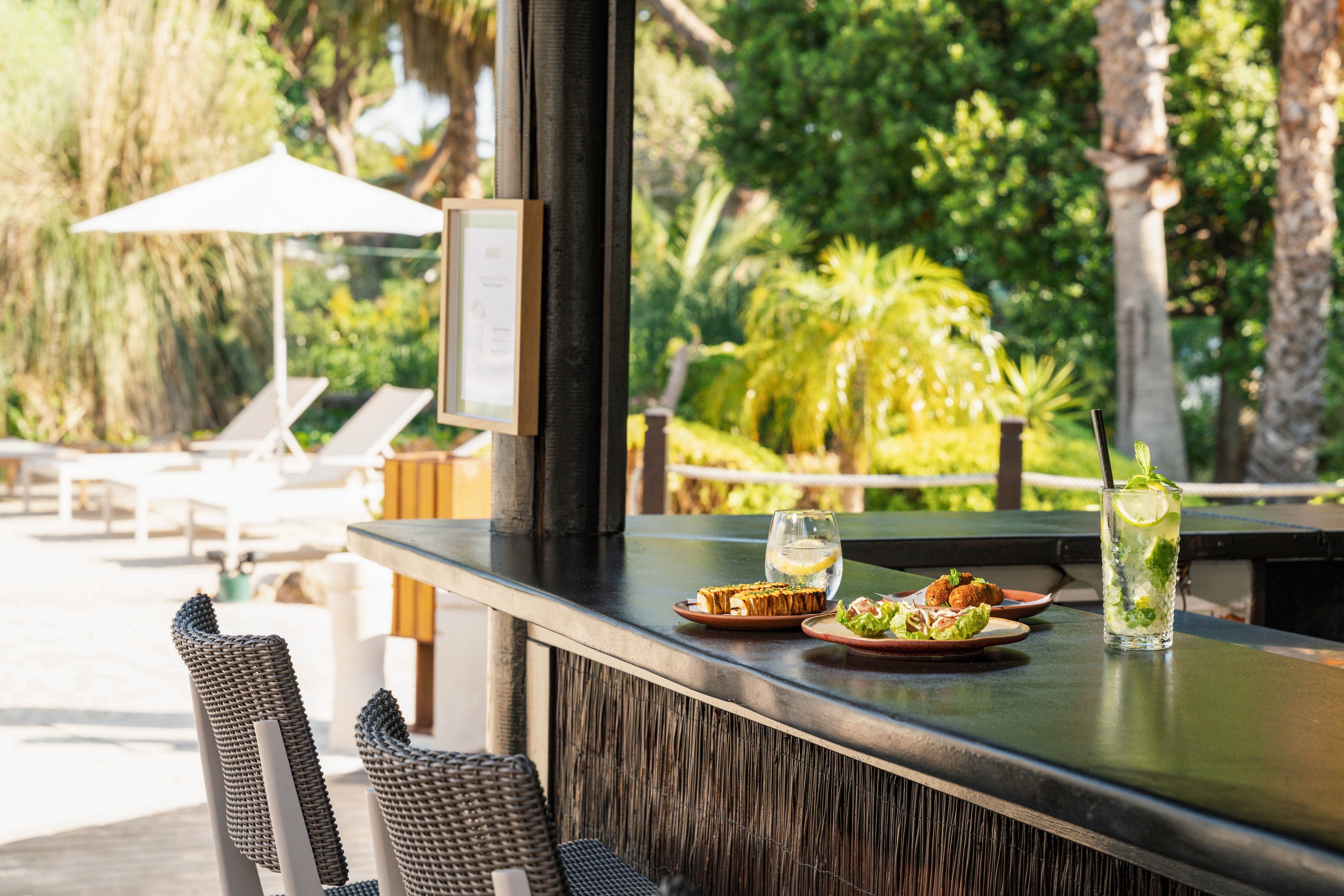An outdoor bar counter displays refreshing cocktails and plates of food in a sunny resort setting. Wicker bar stools line the counter, facing lush green palm trees and landscaped gardens. In the background, white lounge chairs and a large umbrella suggest a relaxing poolside or terrace area. The bright daylight and greenery create a relaxed, vacation atmosphere.