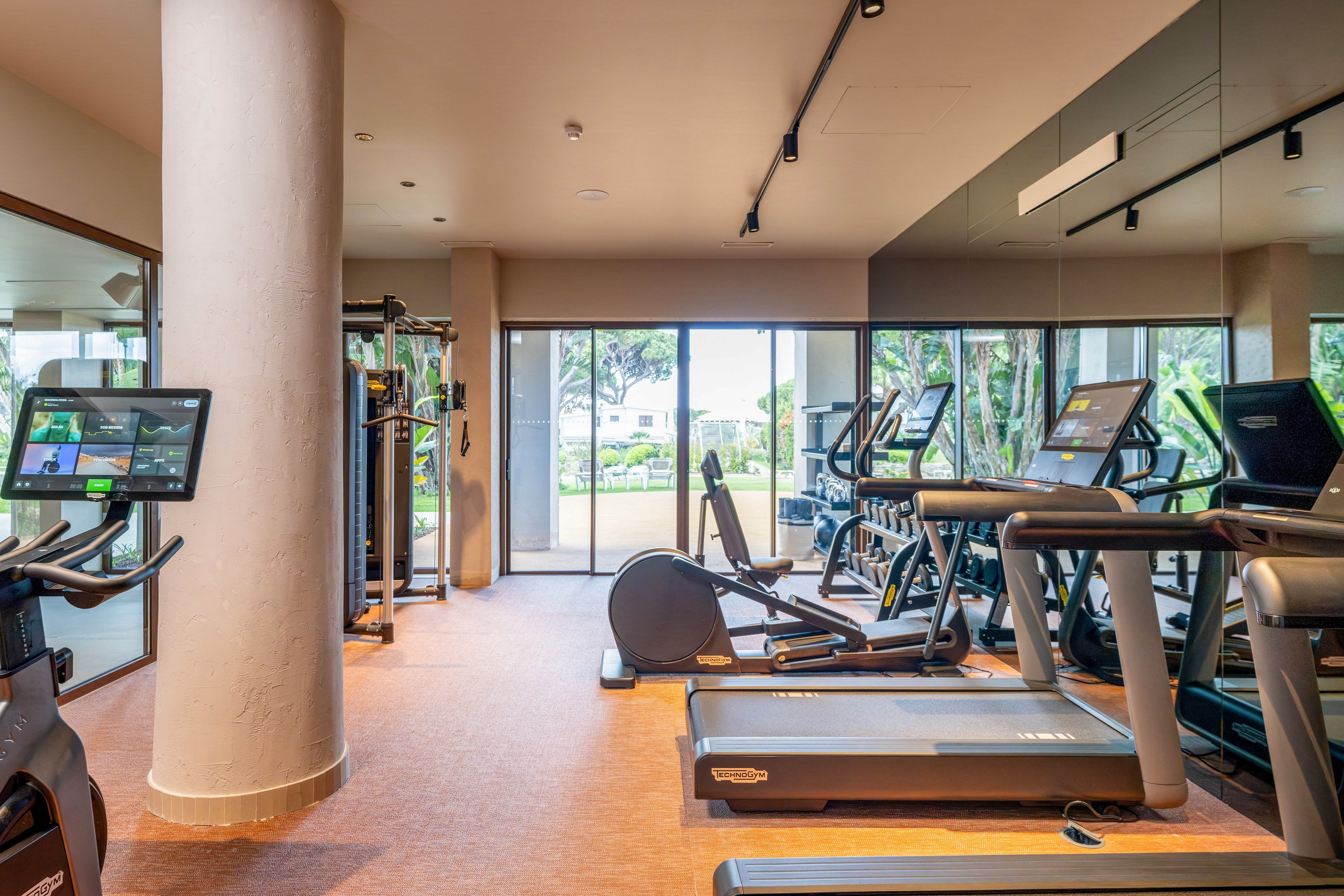A bright, modern gym features several treadmills and elliptical machines arranged in front of large windows. The space includes digital screens on the equipment and mirrors along one wall, creating a spacious feel. Natural light enters through glass doors, revealing greenery and an outdoor area beyond. The clean, organized environment suggests a contemporary hotel or residential fitness center focused on cardio workouts.