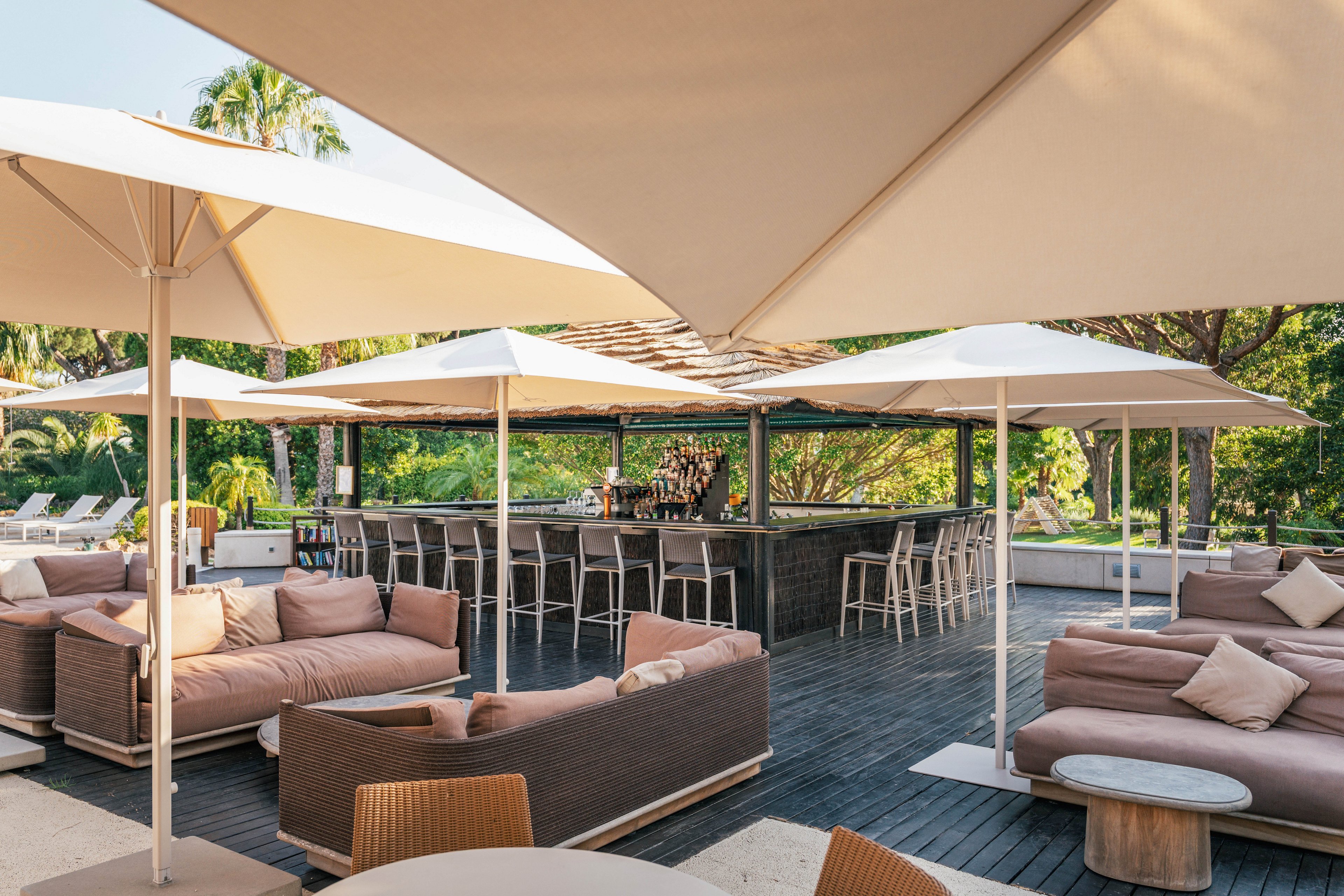 An open-air bar lounge features cushioned sofas and armchairs arranged around a central bar counter. Large white sun umbrellas provide shade over the seating area on a dark wooden deck. The setting is surrounded by lush green trees and palm trees, creating a relaxed vacation atmosphere. Neutral-toned furniture and soft pillows contribute to a modern, comfortable outdoor design.