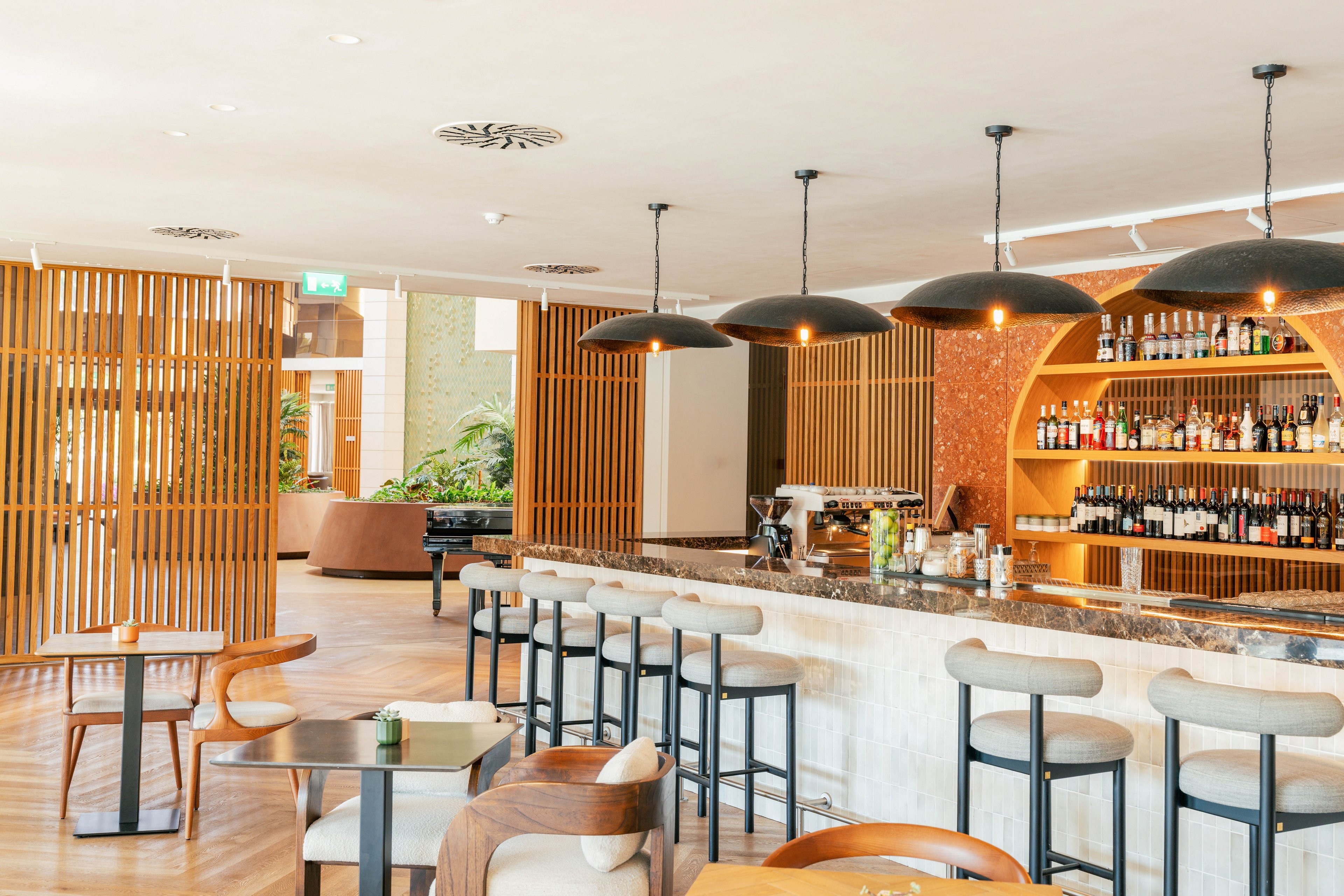 Bright contemporary bar area features a long marble counter with high stools and a well-stocked liquor display. Wooden slatted partitions and curved shelving create a warm, stylish interior design. Pendant lights hang above the bar, while small tables and chairs provide additional seating in the foreground. Natural light and indoor plants contribute to an inviting, relaxed atmosphere.