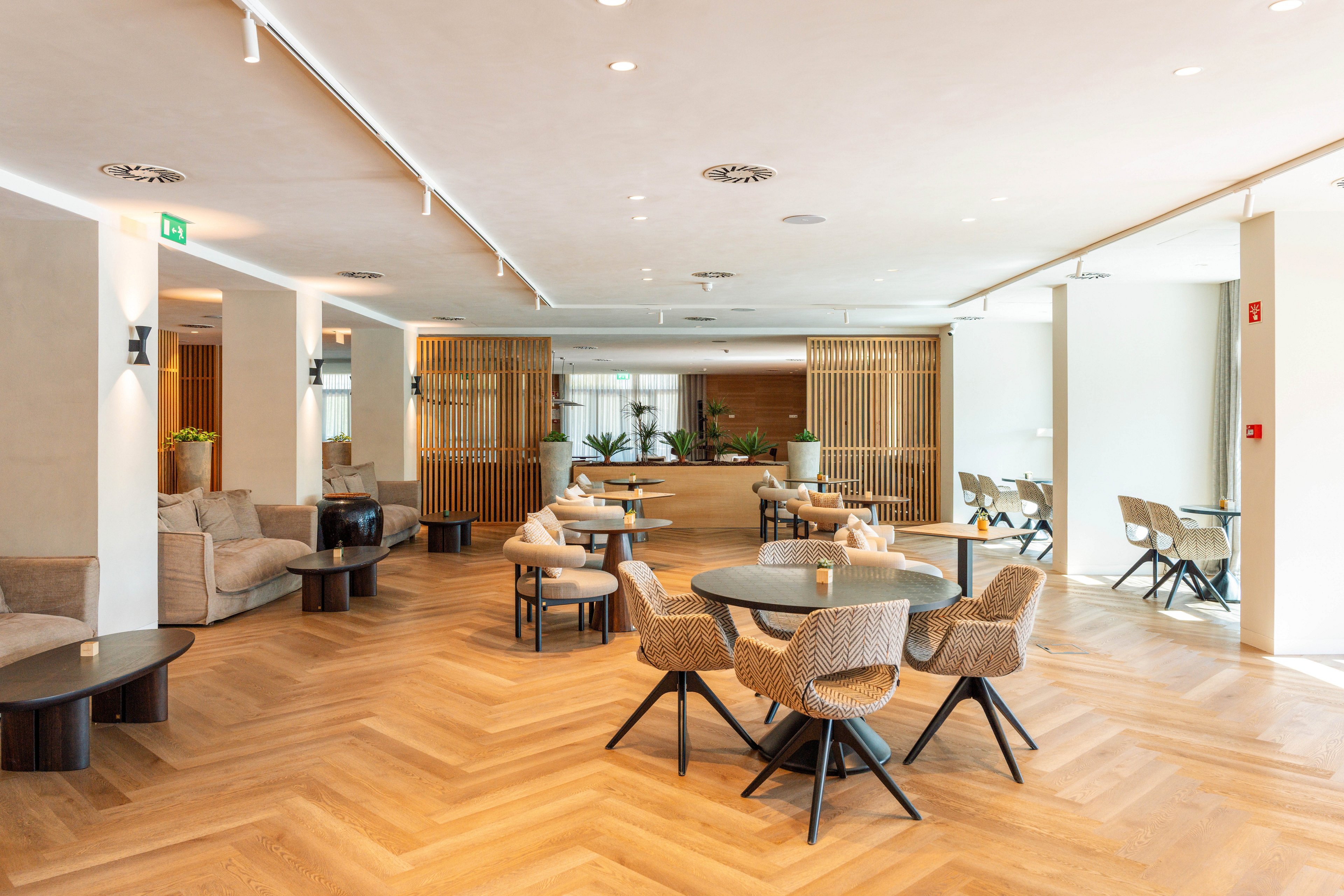 Spacious contemporary lounge with round tables and woven chairs is shown in a bright interior. Light wood herringbone flooring extends across the room, complemented by neutral-toned sofas and side tables. Vertical wooden slat partitions and indoor plants add warmth and texture to the minimalist design. Large windows in the background bring in natural light, creating an inviting and relaxed atmosphere.