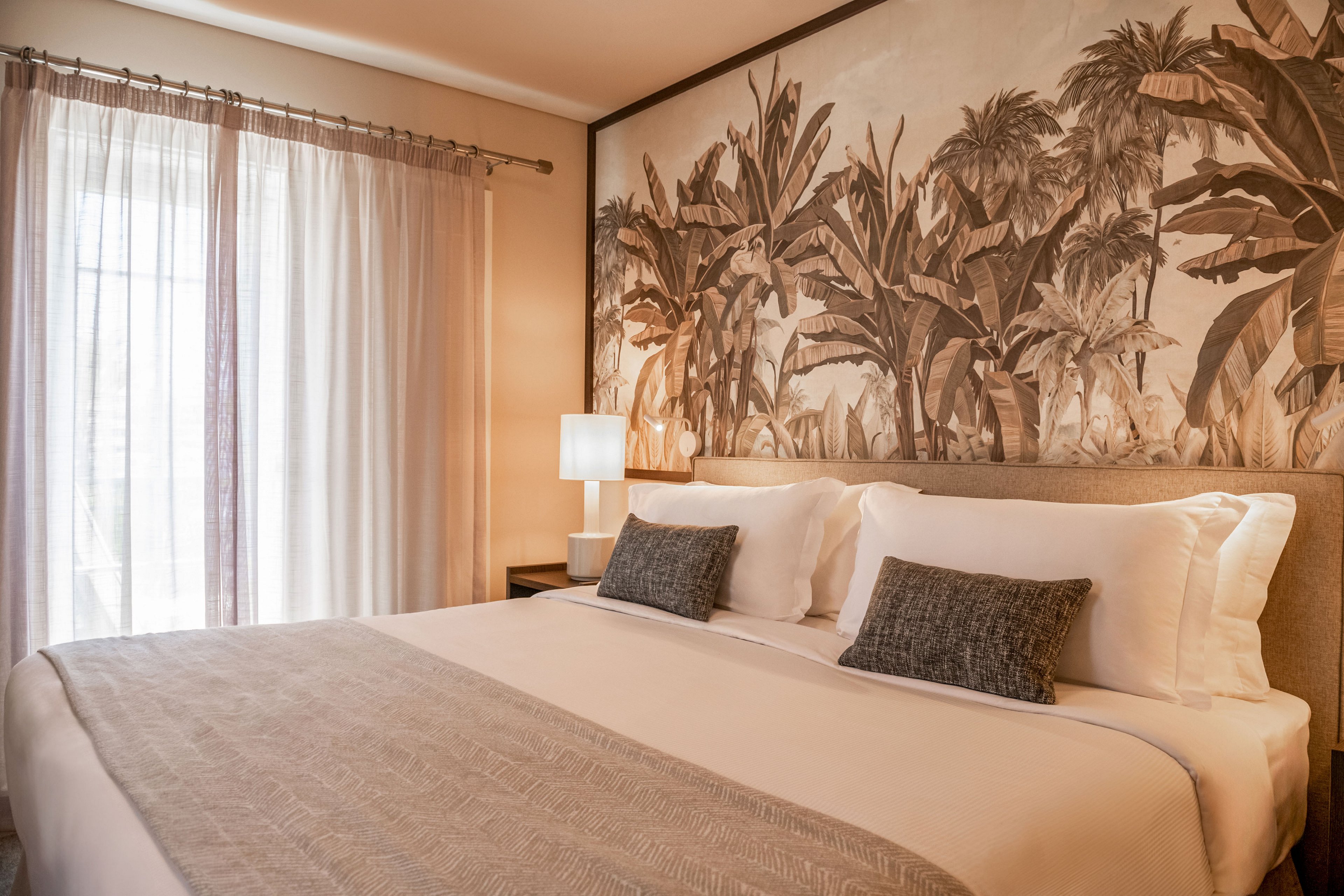 A neatly made bed with white linens and neutral pillows is the focal point of this cozy bedroom. Behind the bed, a large wall mural features tropical foliage in warm sepia tones, adding visual interest. Soft natural light filters through sheer curtains beside the bed, complemented by a glowing bedside lamp. The overall setting feels calm and inviting, with a warm color palette and minimal decor.