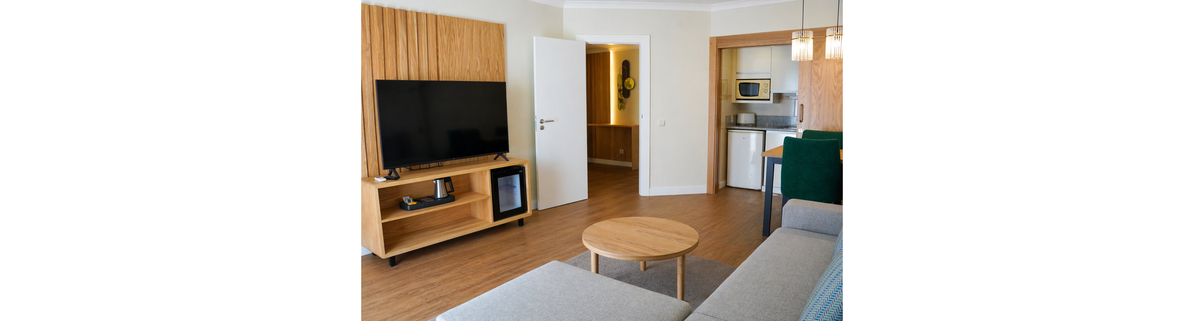 A contemporary living room features a large flat-screen television on a wooden media console and a small refrigerator beneath. A light gray sofa and round wooden coffee table sit on a warm wood floor, creating a cozy seating area. An open doorway leads to a hallway and a compact kitchenette with a microwave, sink, and cabinets. Pendant lights hang above a small dining table with green upholstered chairs, adding a modern touch to the neutral interior.