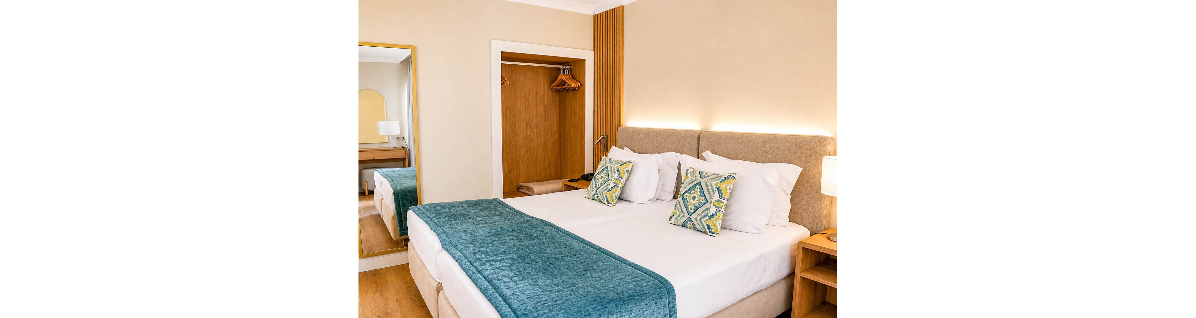 A neatly made double bed with white linens and teal throws is the focal point of this modern bedroom. Patterned accent pillows add color against a neutral upholstered headboard with soft backlighting. An open wooden wardrobe with hangers and a bedside table frame the bed area. A wall mirror reflects part of the room, enhancing the sense of space and light.