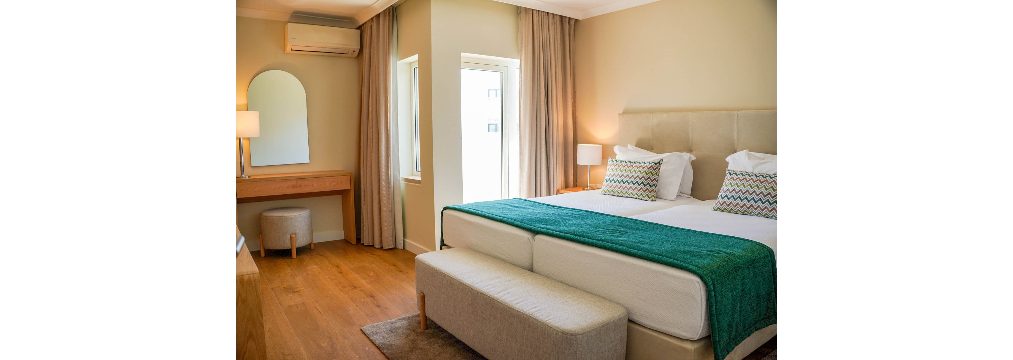 A contemporary hotel bedroom features a large double bed with a padded headboard and a teal throw blanket. Two patterned pillows add color against neutral bedding and soft beige walls. Floor-to-ceiling curtains frame tall windows, allowing natural light into the room. A wooden vanity with a mirror, stool, and table lamp sits along one wall, creating a cozy and inviting atmosphere.