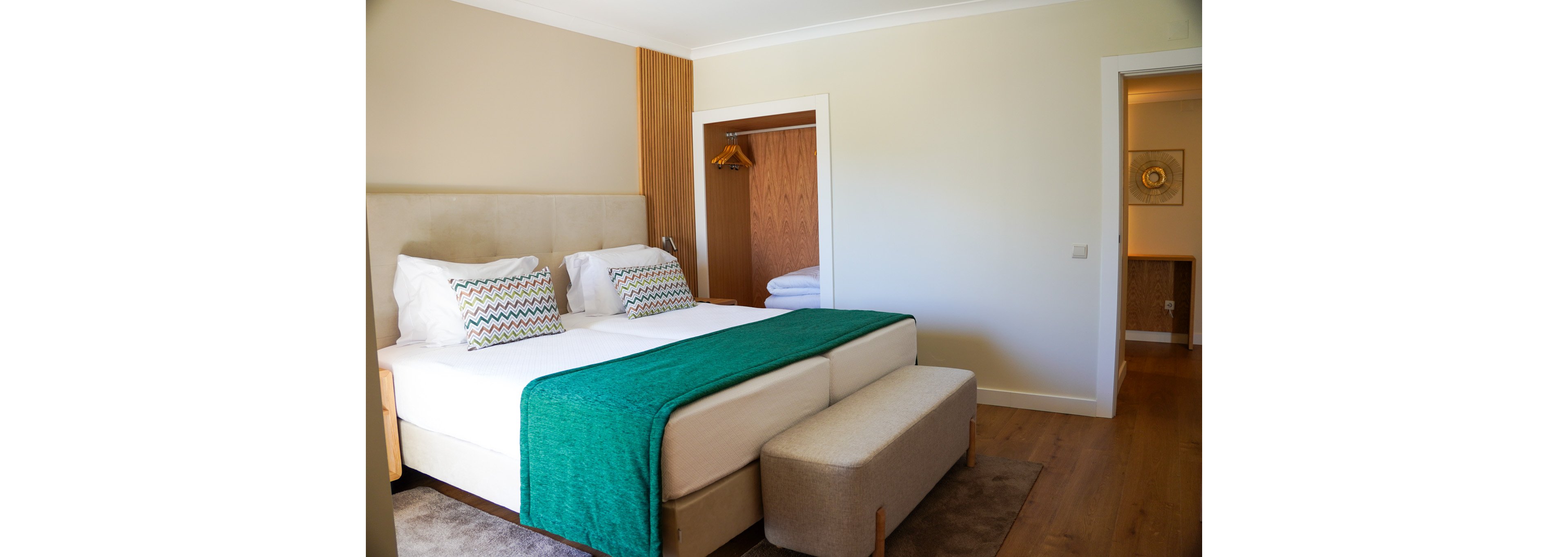 A contemporary hotel bedroom features a large double bed with white linens and teal bed runner. Two patterned pillows add color against a neutral upholstered headboard. An open wardrobe with wooden hangers and folded towels is visible beside the bed. A small upholstered bench sits at the foot of the bed, and a hallway leads to another softly lit area, creating a calm and inviting atmosphere.