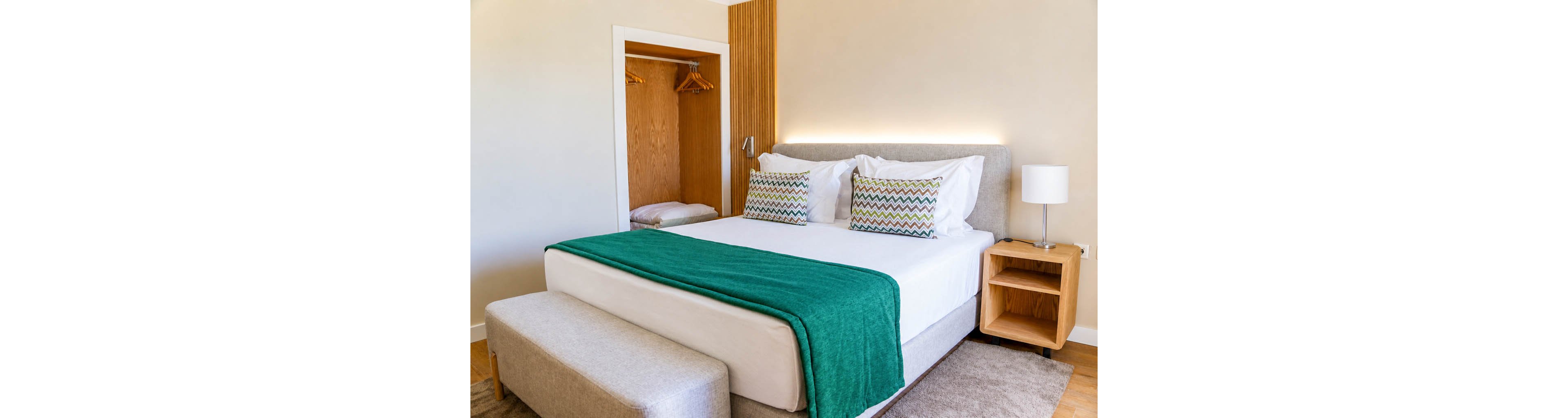 A contemporary bedroom features a large neatly made bed with white linens and a teal throw blanket. Two patterned pillows add color and texture to the headboard area, which is softly lit from behind. An open wooden closet with hangers and a folded blanket is visible beside the bed. A wooden nightstand with a white table lamp and a cushioned bench at the foot of the bed complete the cozy, minimalist space.