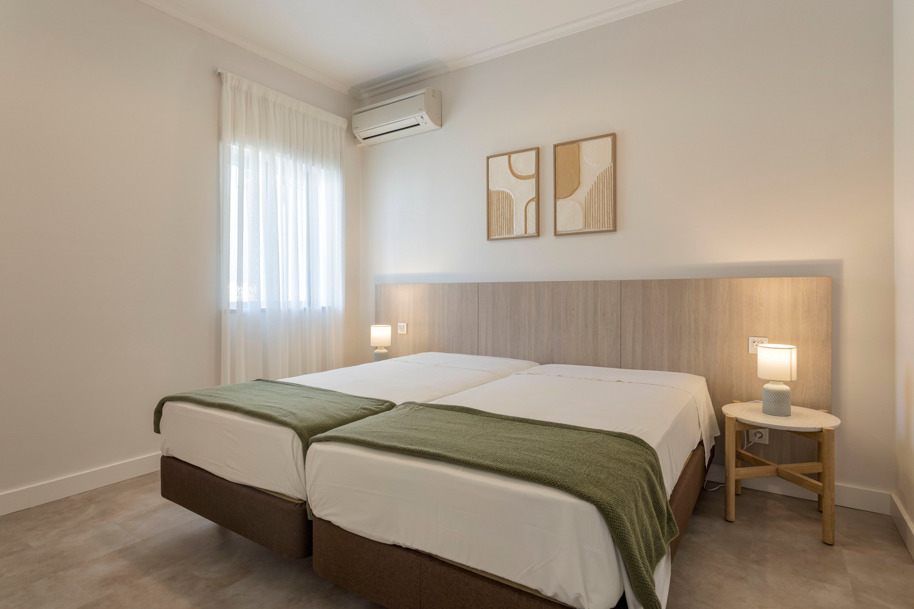 Bright, modern bedroom featuring two neatly made twin beds with white linens and green throws. The room has light neutral walls, a wooden headboard, and a small bedside table with a lit lamp. Sheer curtains cover the window, allowing soft natural light to enter. An air conditioning unit and abstract wall art complete the clean, minimalist decor.