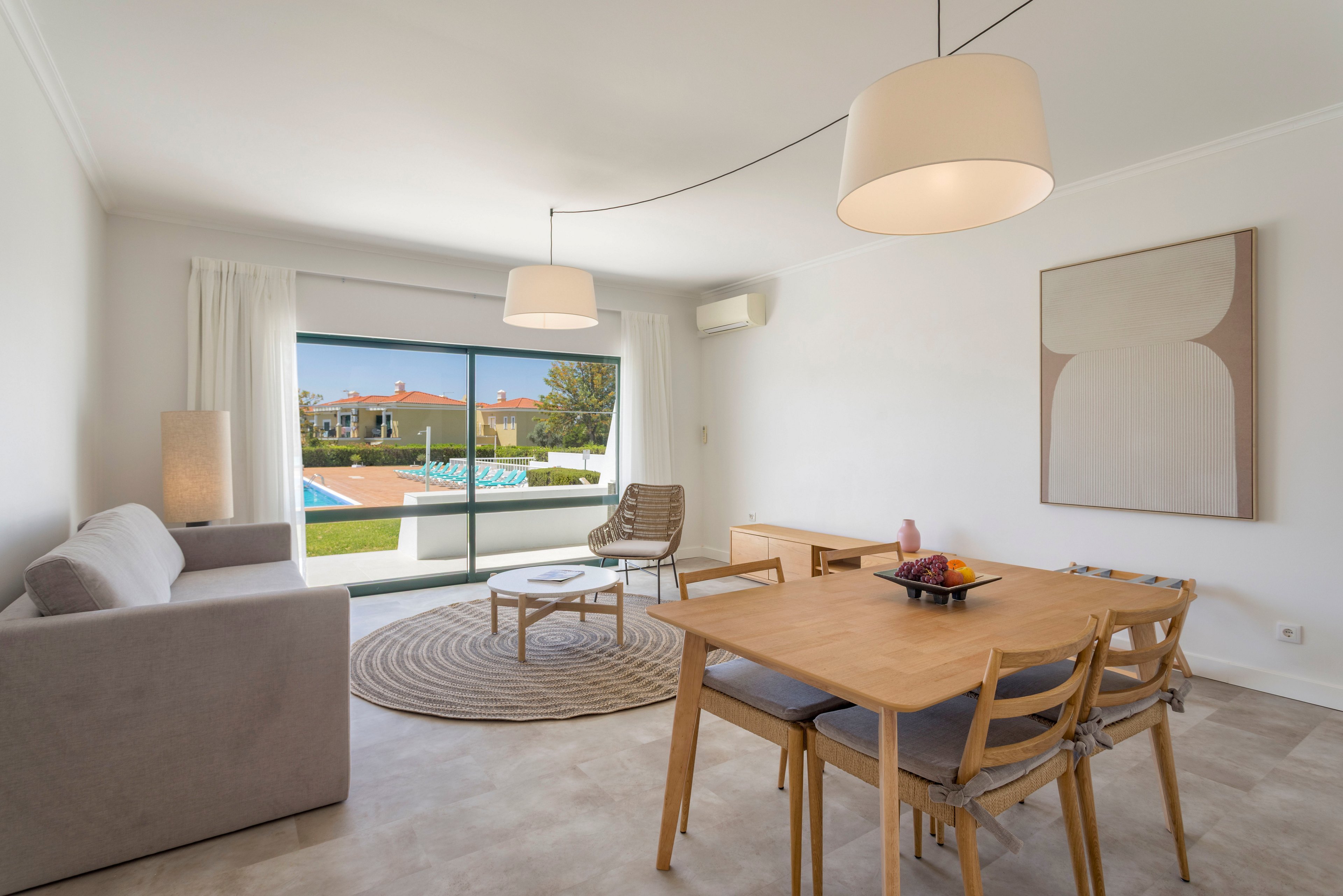 Contemporary living and dining area features a neutral-toned sofa, wooden table, and minimalist decor. Large sliding windows open to a sunny garden with a swimming pool and residential buildings in the background. Soft pendant lighting, a round rug, and abstract wall art create a clean, modern aesthetic. The space feels airy and inviting, ideal for relaxing or entertaining.