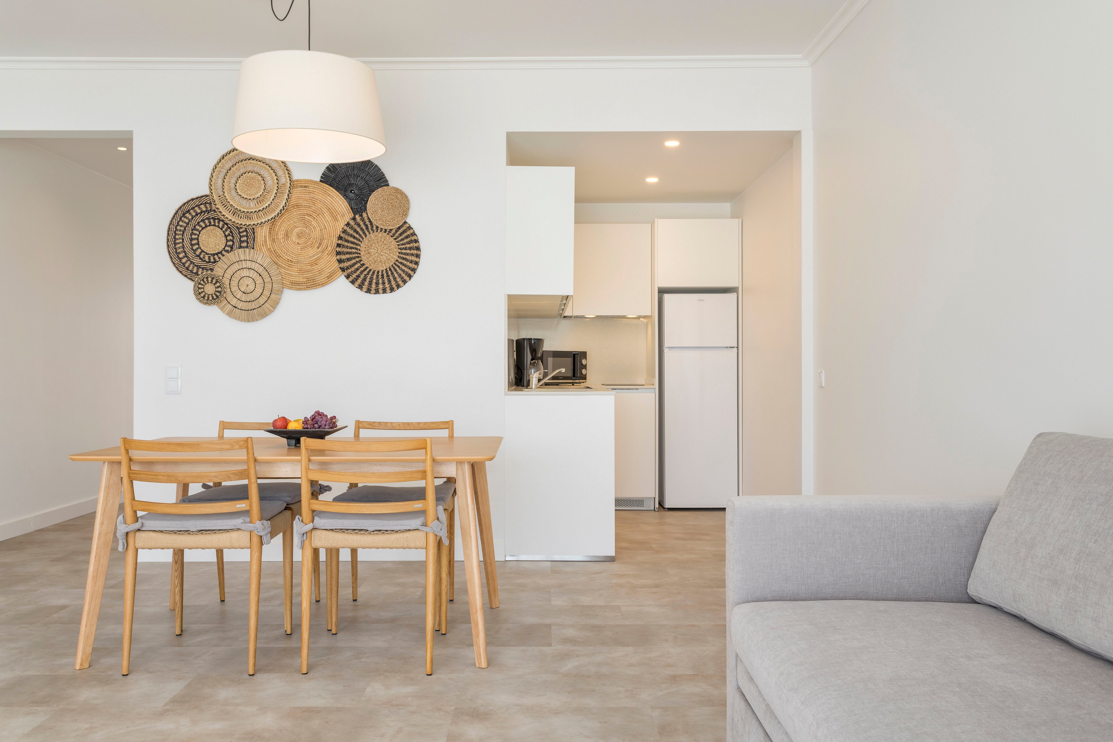 A bright minimalist dining area features a wooden table with four matching chairs and a decorative wall installation of circular woven patterns. In the background, a compact white kitchen includes cabinetry, a refrigerator, and small appliances on the countertop. A large pendant light hangs above the table, illuminating a bowl of fruit. On the right side, a light gray sofa suggests an open-plan living and dining space with neutral tones and clean lines.