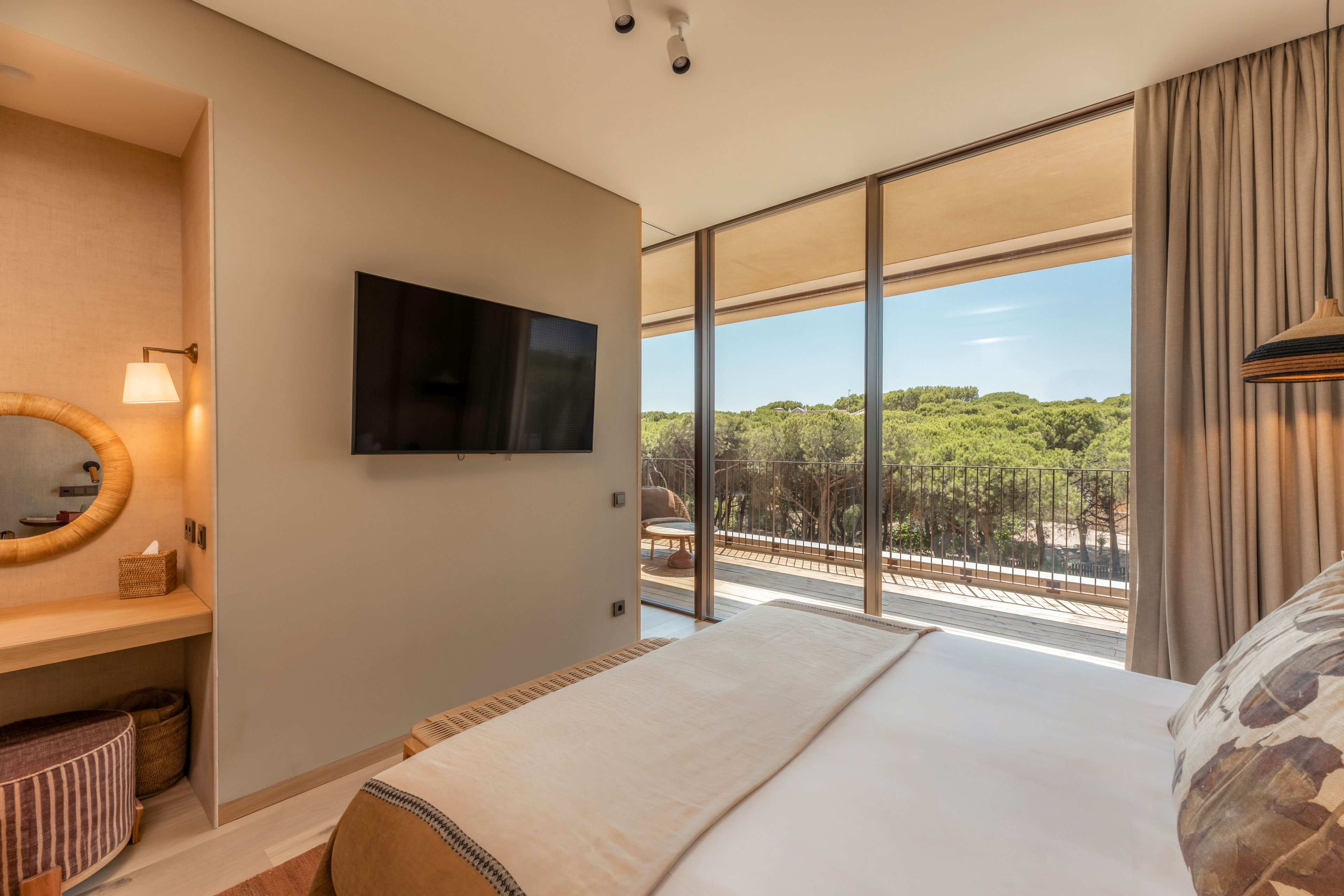 Contemporary hotel bedroom featuring a large bed and wall-mounted flat-screen TV. Floor-to-ceiling windows open to a balcony overlooking a lush green forest landscape. A cozy vanity area with a round mirror and warm lighting sits to the side, complemented by neutral tones and soft textiles. The overall atmosphere is calm, inviting, and filled with natural light.