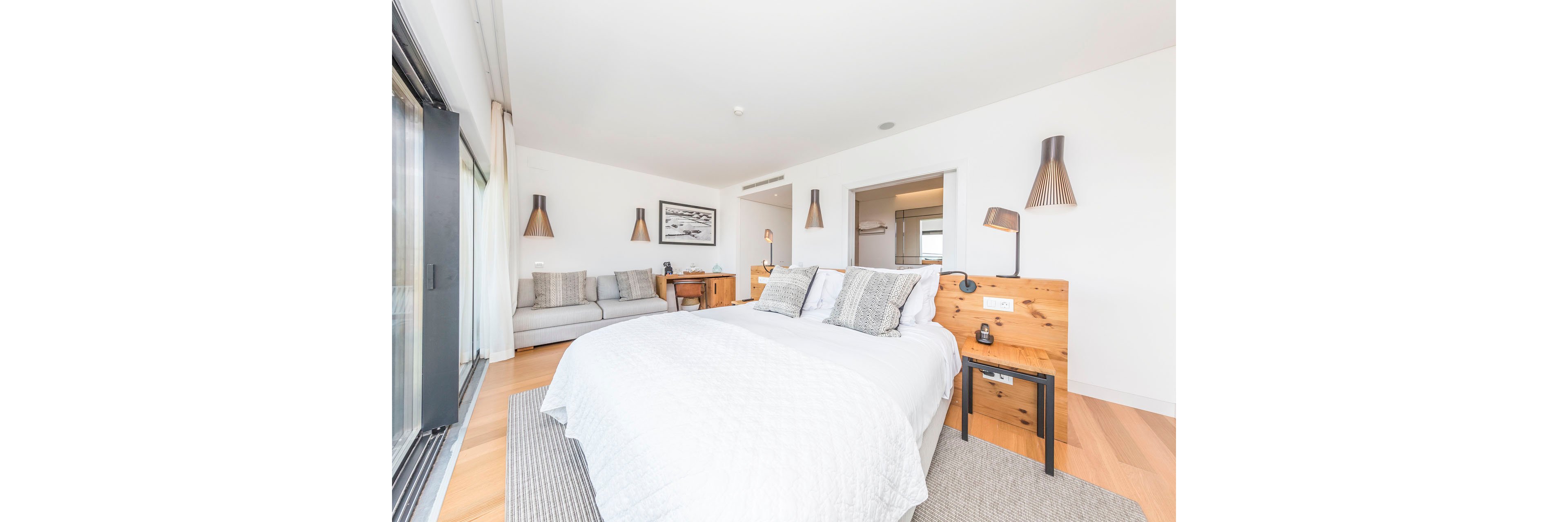 Spacious contemporary bedroom featuring a large bed with white bedding and textured pillows. A light gray sofa with cushions sits against the wall, creating a comfortable seating area. Wooden bedside tables and wall-mounted lamps add warmth to the minimalist decor. Large windows bring in abundant natural light, highlighting the clean lines and neutral color palette.