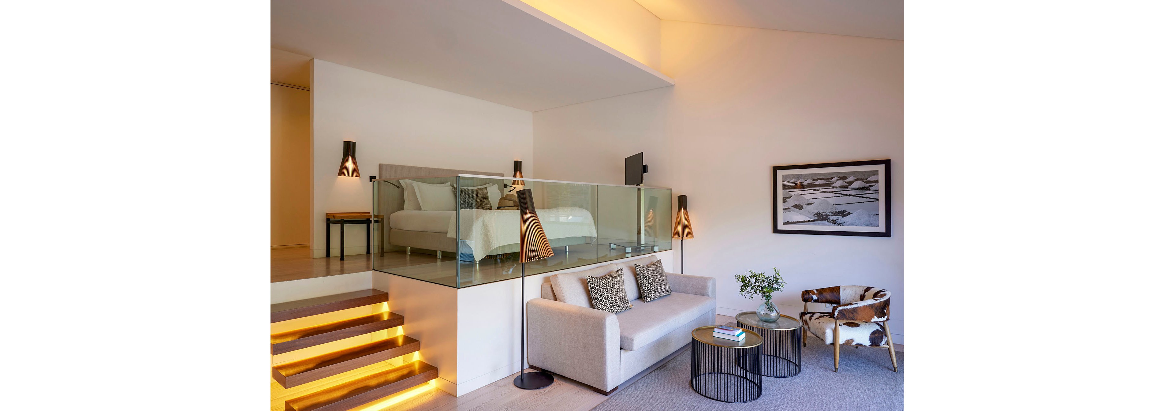 Contemporary split-level suite features a raised bedroom area with a double bed behind a glass railing. A cozy living space below includes a light gray sofa, patterned armchair, and round side tables. Warm accent lighting illuminates the staircase and pendant lamps, creating an inviting atmosphere. Minimalist decor, neutral tones, and a framed wall photograph emphasize modern, upscale comfort.